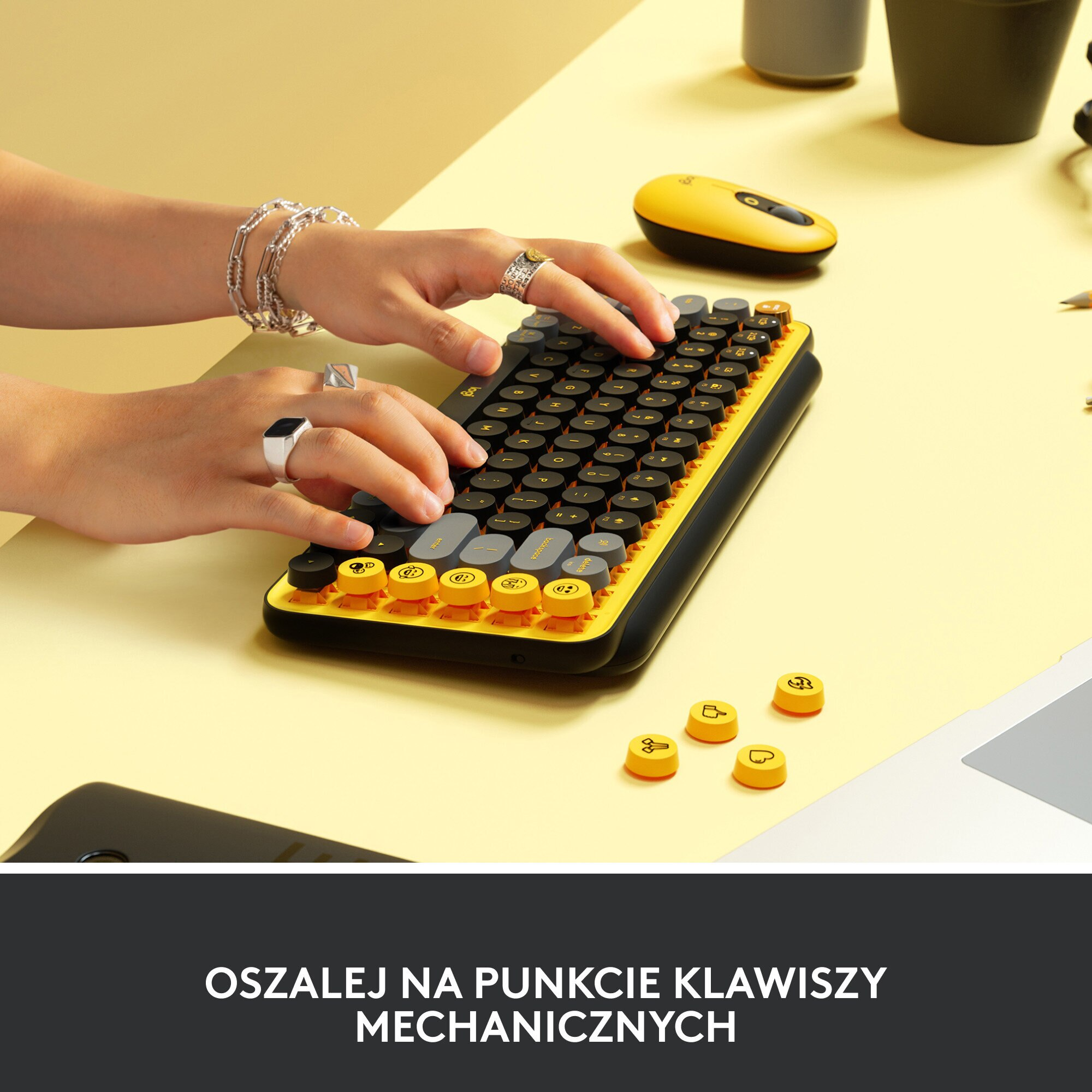 Dłonie osoby na żółto-czarnej klawiaturze z okrągłymi klawiszami. Różne akcesoria w tle.
