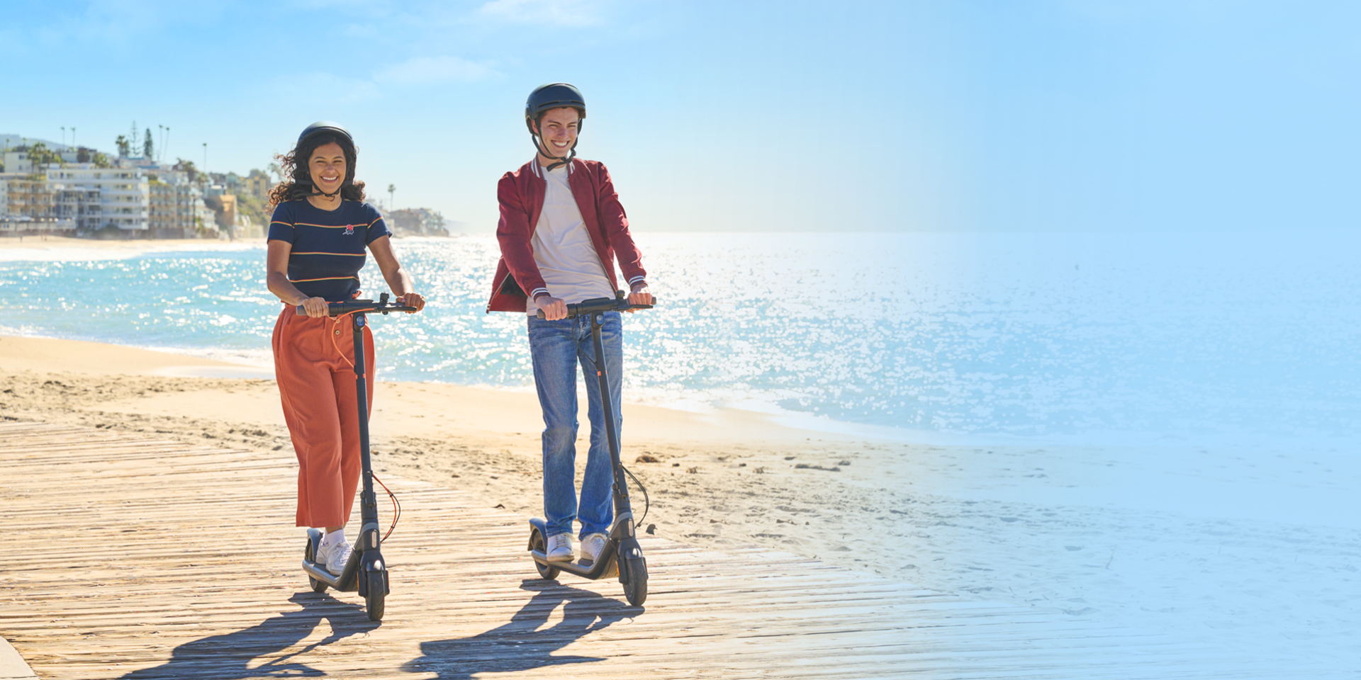 Két ember elektromos robogóval egy fa pallón a tengerparton, kék égbolttal.