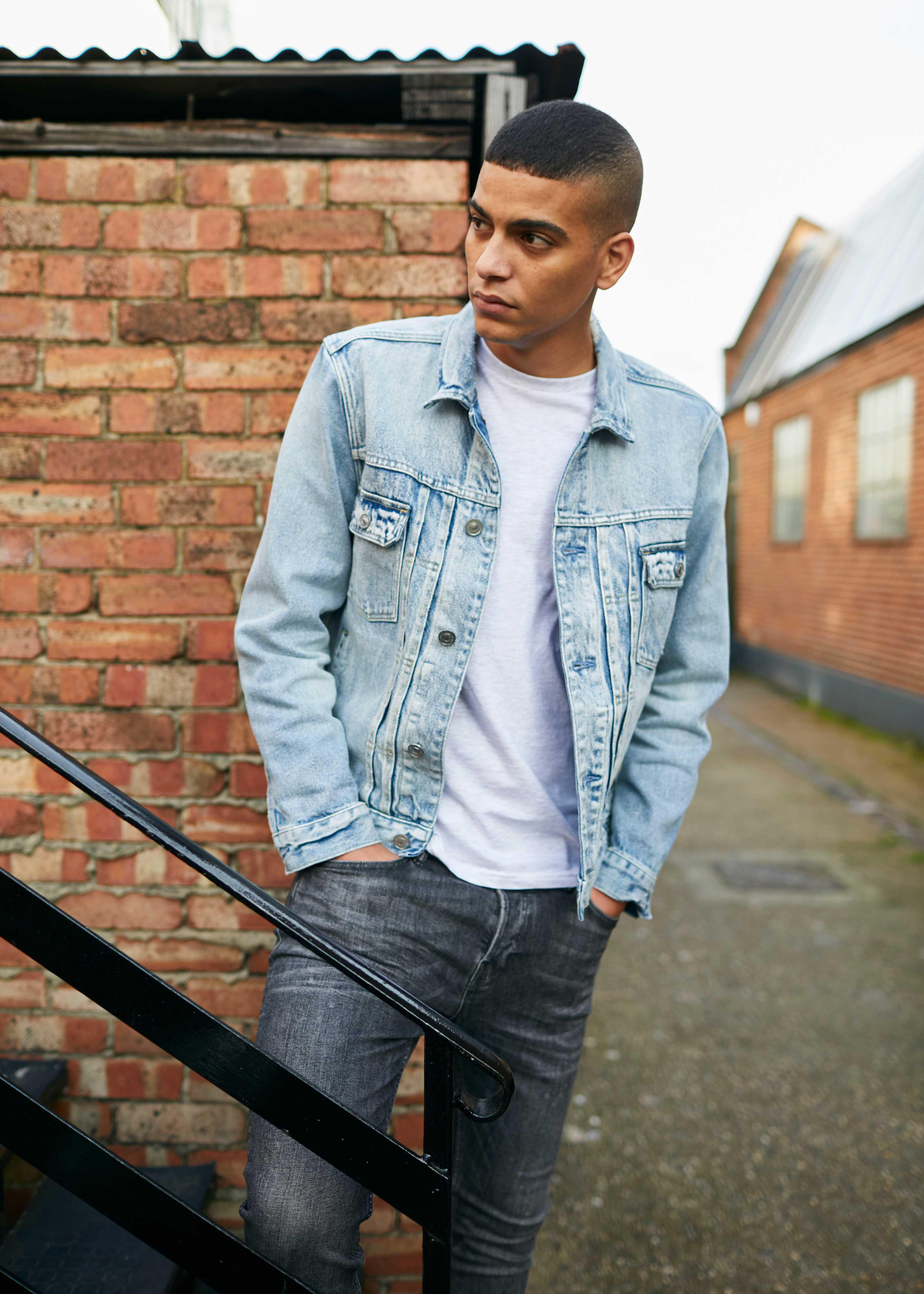 Junger Mann in hellblauer Jeansjacke und grauer Jeans, lehnt an einer Backsteinmauer.
