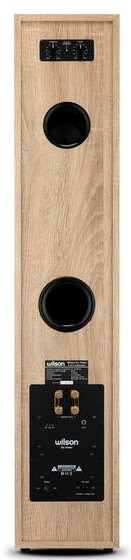 Widok z tyłu wysokiego głośnika z jasnego drewna z dwoma czarnymi otworami głośnikowymi i czarnym panelem.