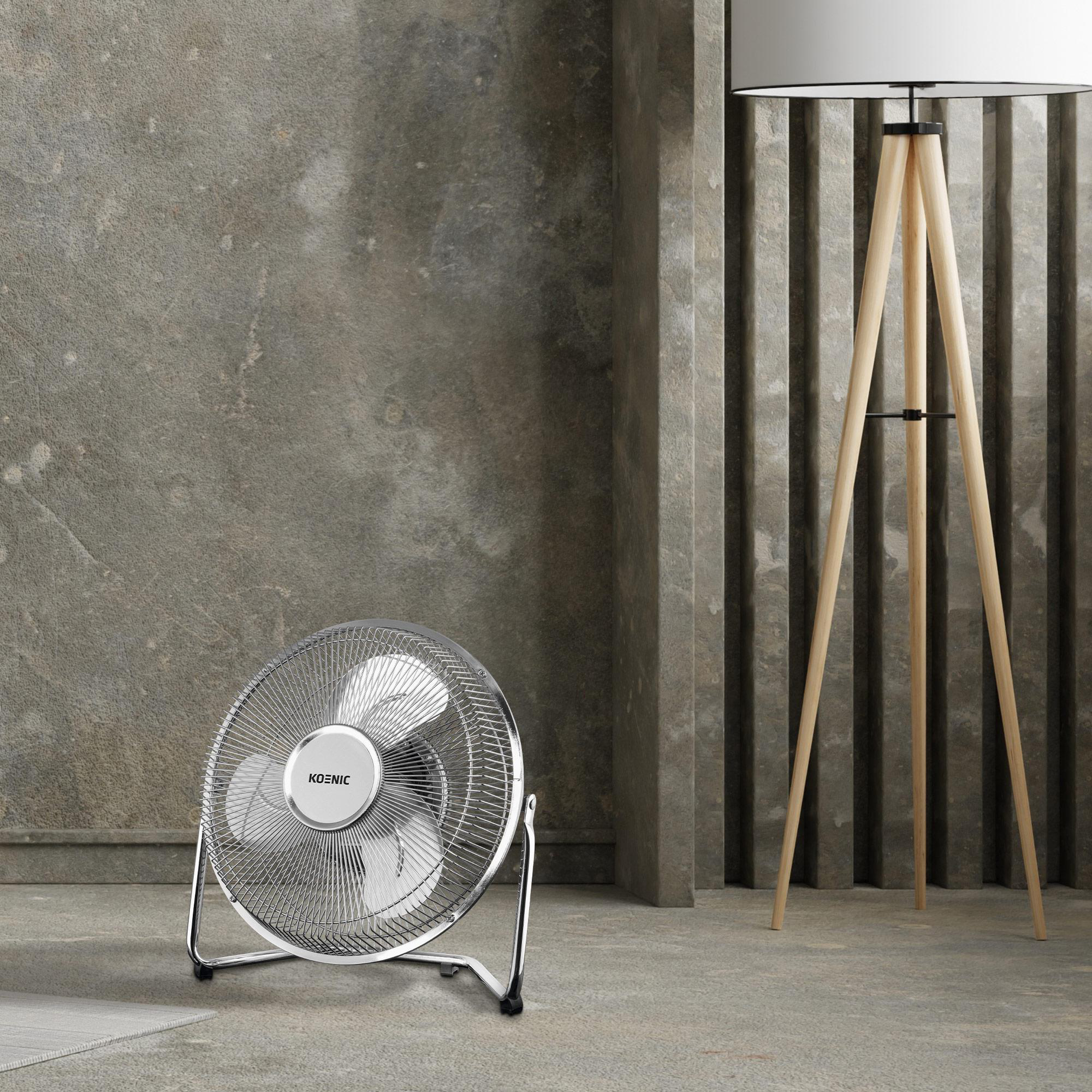 Ein silberner Ventilator der Marke "KOENIC" steht auf einem grauen Boden, im Hintergrund eine Holzlampe.