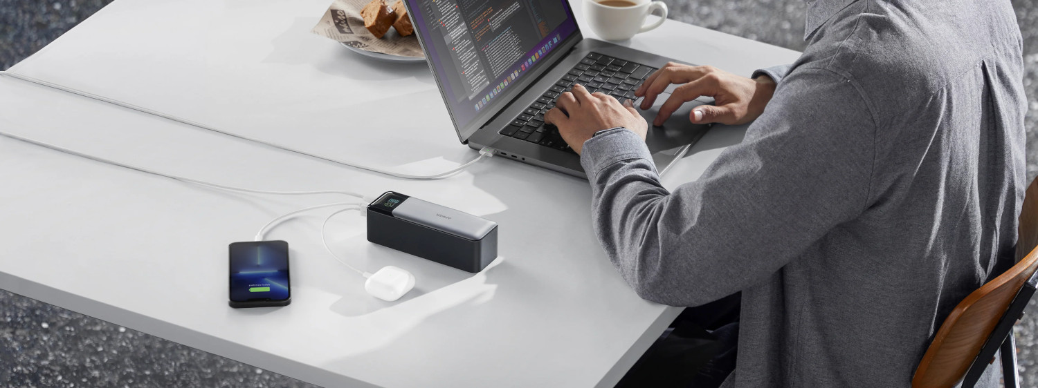 A person working on a laptop connected to a power bank and a phone. A cup of coffee is on the table.
