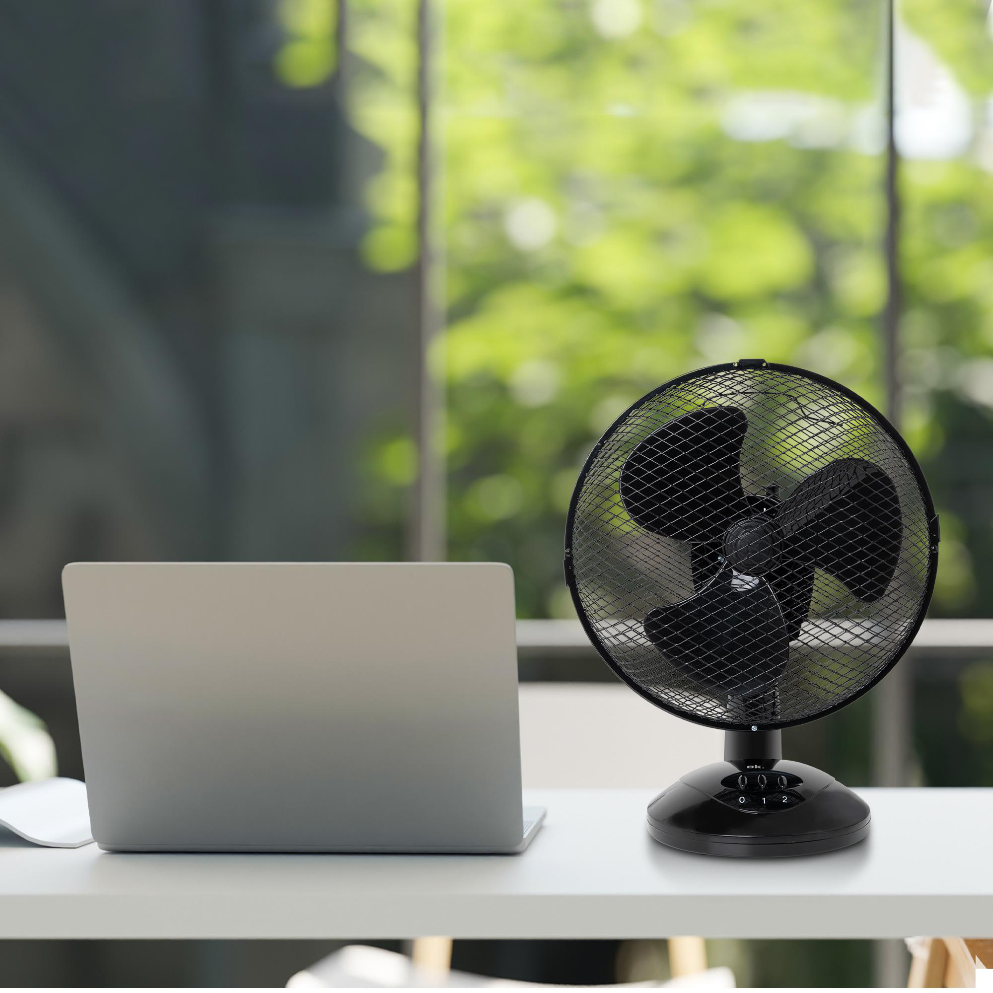 Schwarzer Ventilator auf Schreibtisch mit Laptop, Fenster und Grün im Hintergrund.