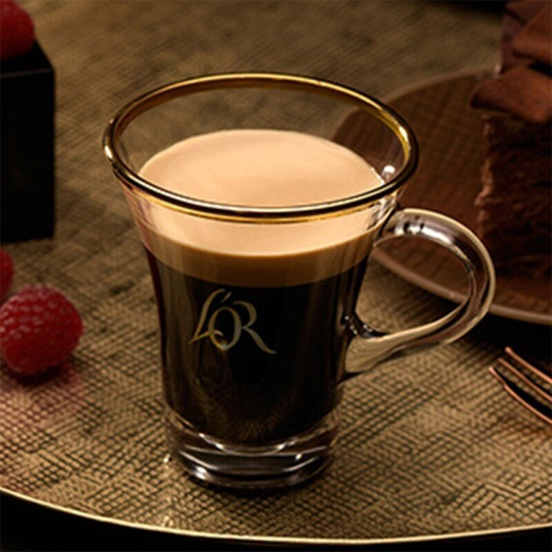 Przezroczysty szklany kubek kawy ze złotą obręczą. Kawa jest warstwowa ze śmietanką i ciemnym espresso. Maliny i ciasto w tle.