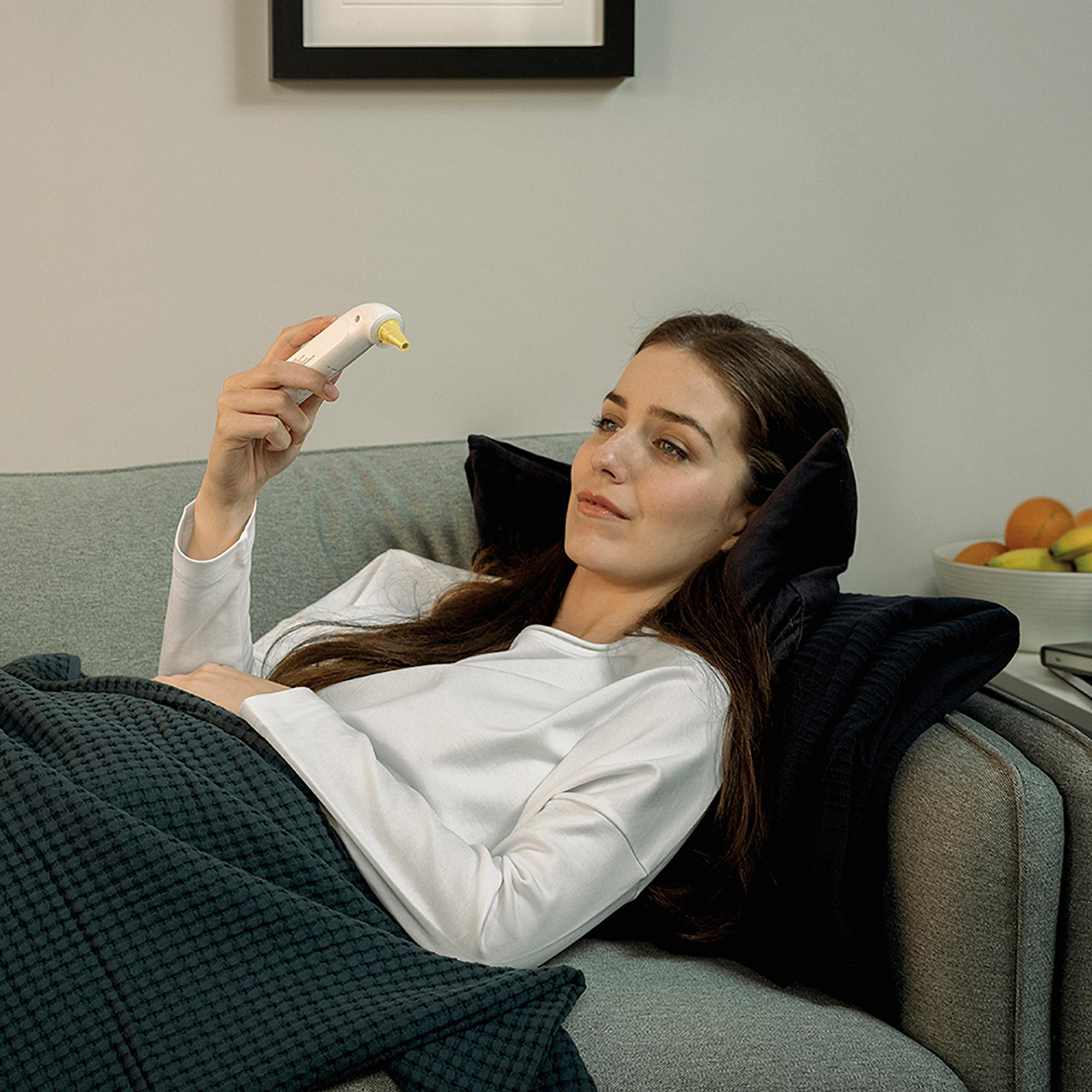 Frau auf grauem Sofa, hält weißes Thermometer. Sie schaut darauf, mit Decke und Kissen zur Unterstützung.