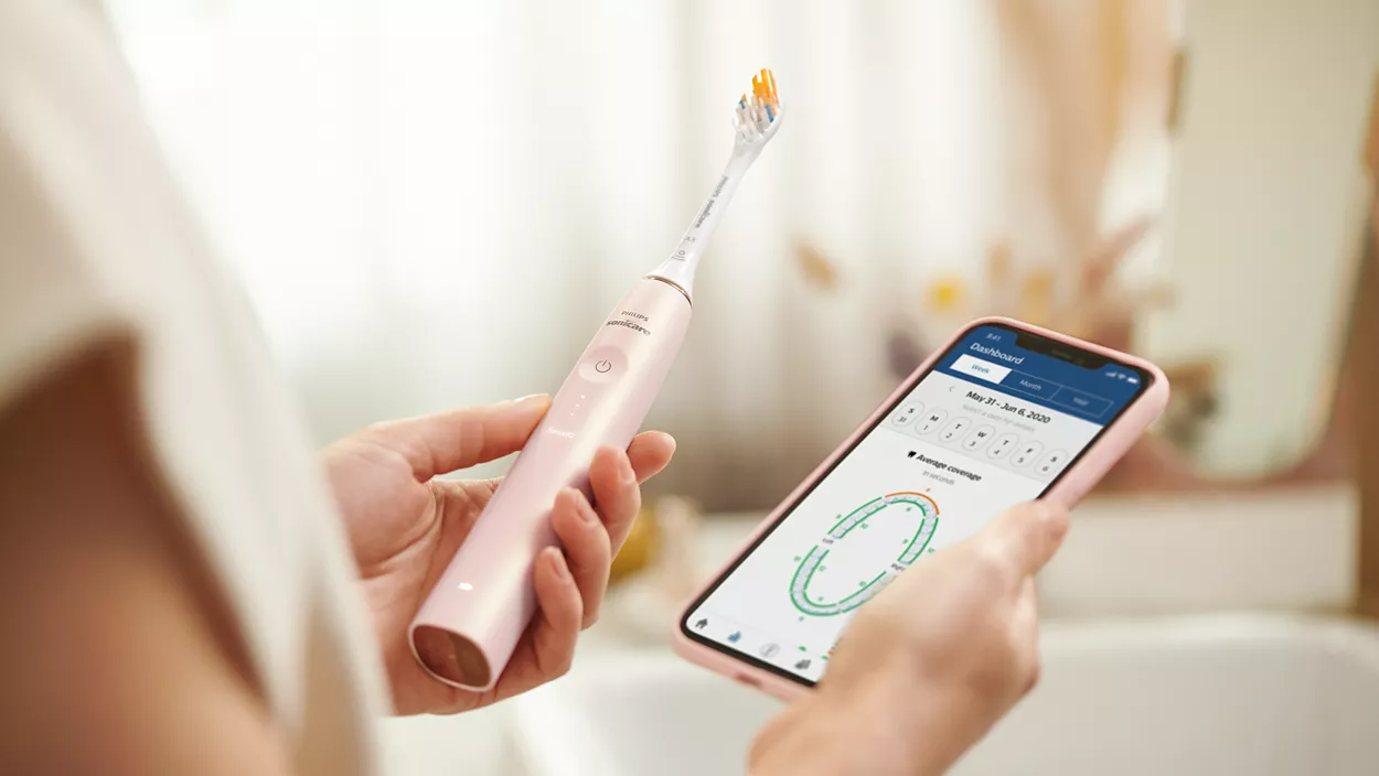 Nő rózsaszín elektromos fogkefét és egy okostelefont tart, ami fogmosási adatokat mutat.