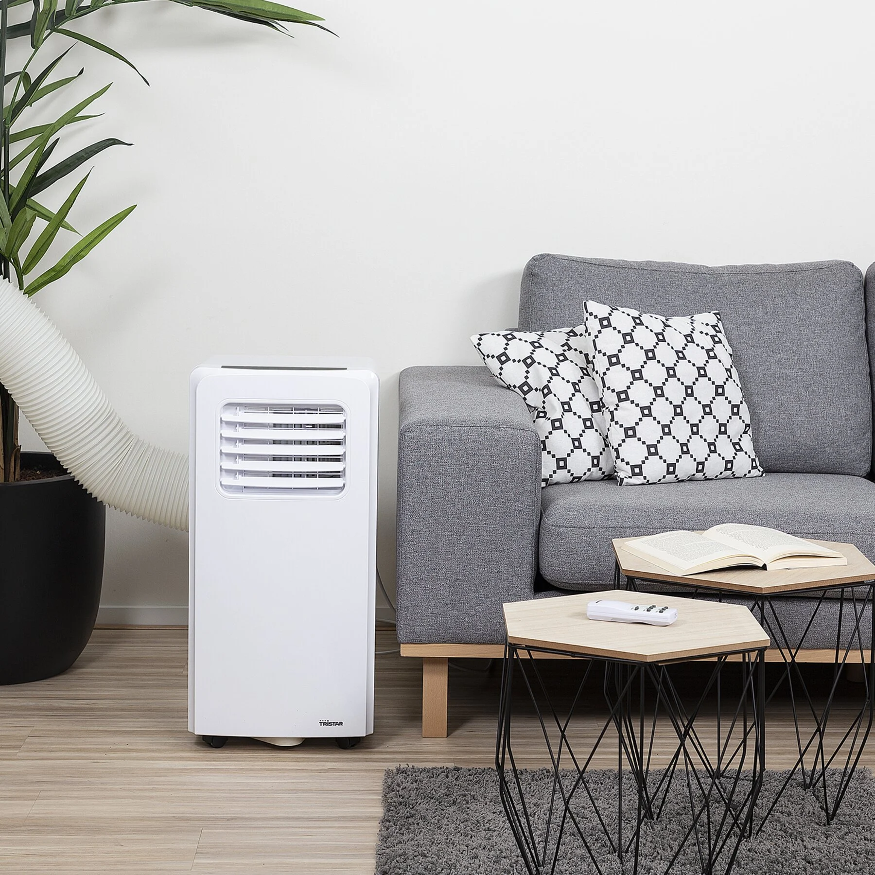 Een witte airconditioner bij een grijze bank met kussens met patronen. Twee tafels, een boek en een afstandsbediening ervoor.