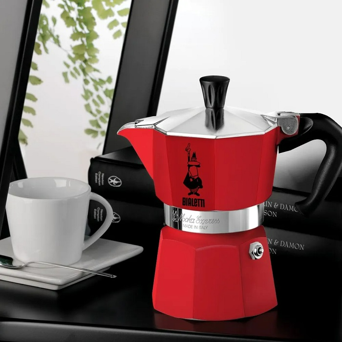 Egy piros Bialetti Moka Express kávéfőző fekete felületen, fehér csészével.