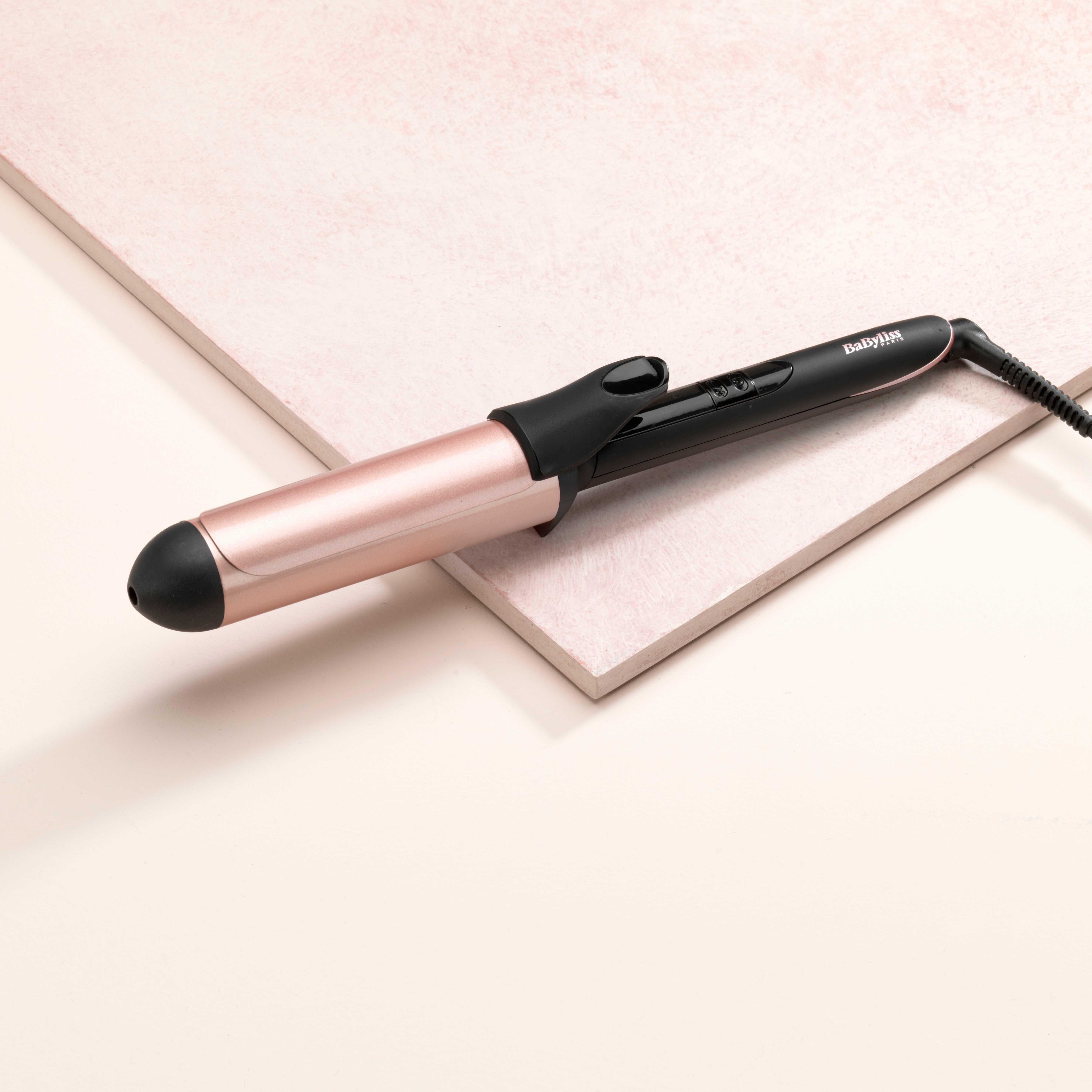 Roségoldener und schwarzer Lockenstab auf einer rosa-weißen Oberfläche. Kabel angeschlossen. Weißer Hintergrund.