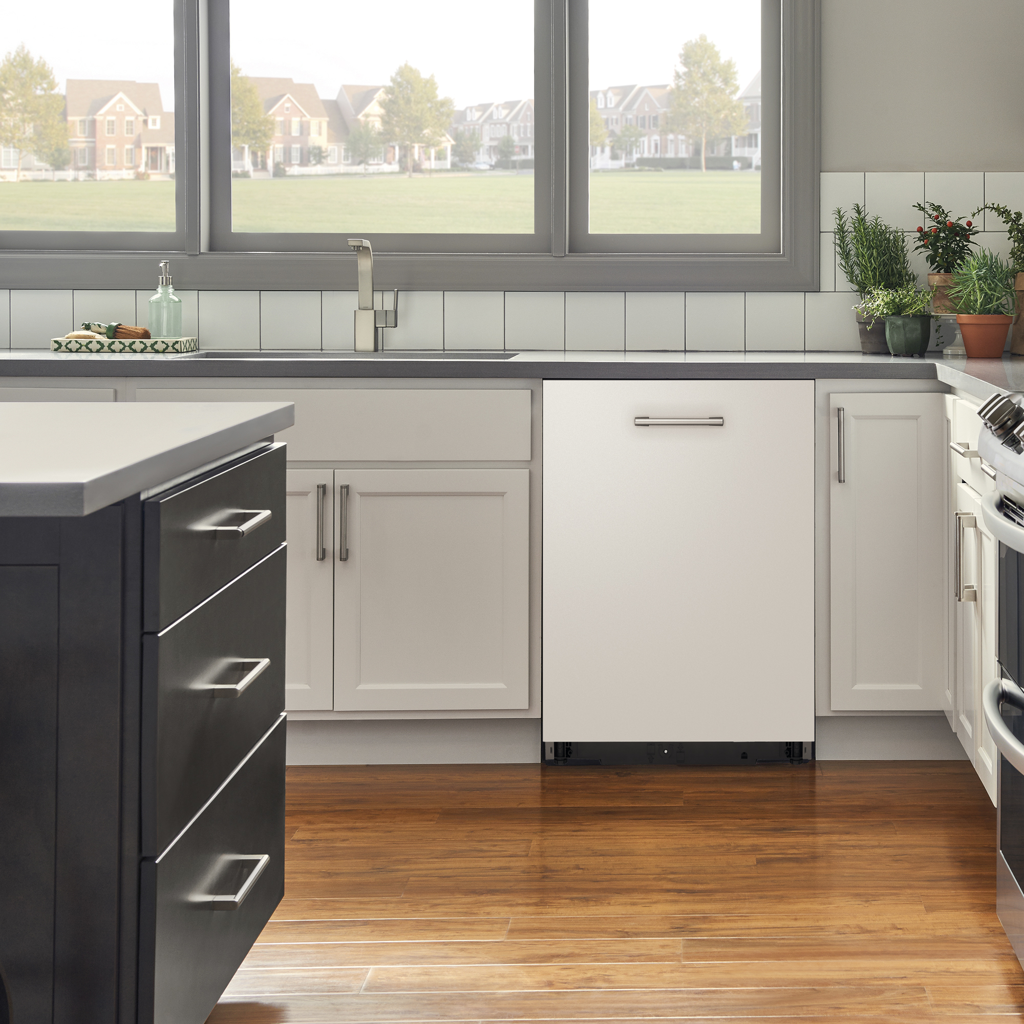 Una cocina con gabinetes blancos, un lavavajillas, pisos de madera y una ventana con vistas a casas.