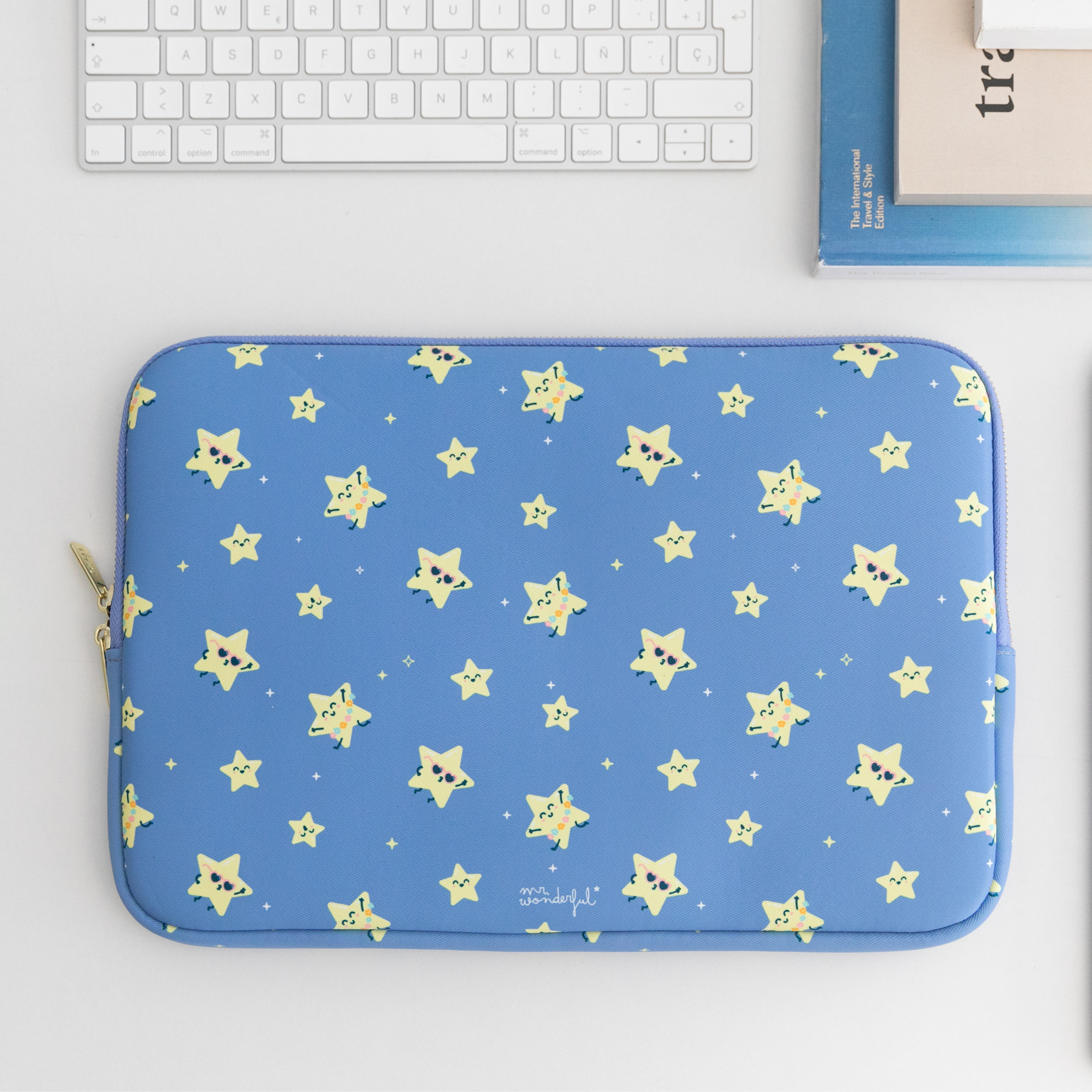 Una funda azul para portátil con patrones de estrellas amarillas sobre un escritorio blanco con teclado y libros.