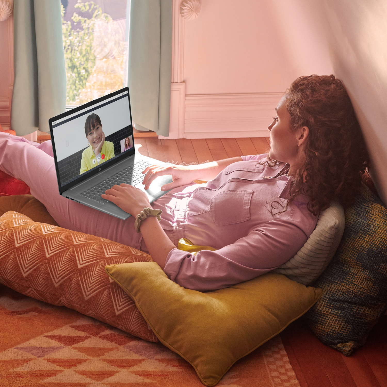 Vrouw in roze top met laptop, videobellen. Kussens en raam op de achtergrond.