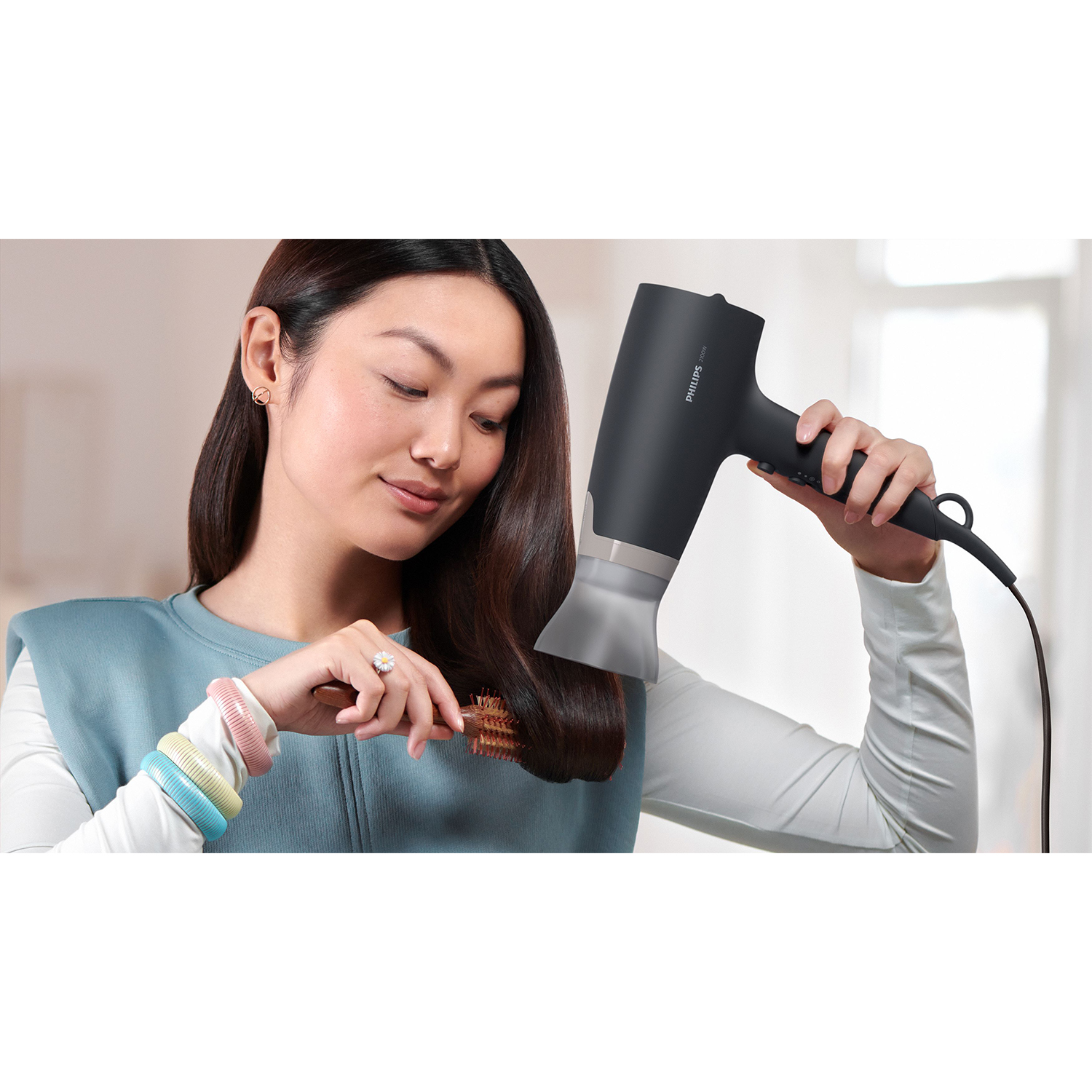 Donna asiatica usa un asciugacapelli nero, guarda in basso. Si spazzola i capelli con una spazzola.