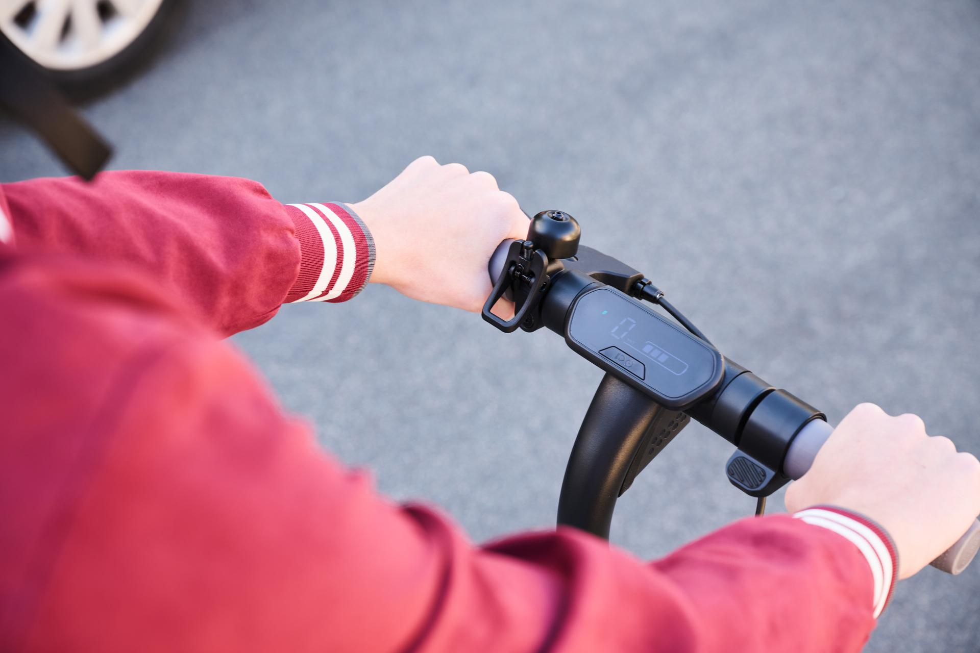Manos de persona en manillar de scooter. El scooter es negro, la persona lleva chaqueta roja.