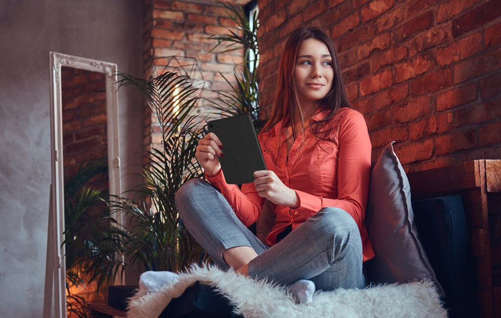 Kobieta w koralowej koszuli siedzi, trzymając tablet. Tło z ceglanej ściany, roślin, lustra i drewnianej ławki.