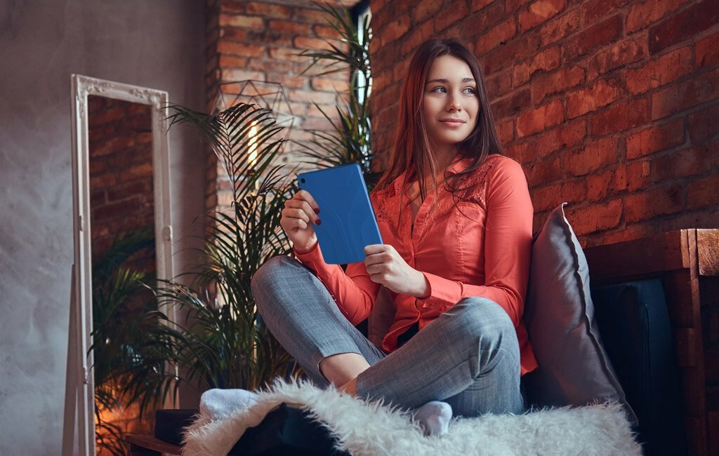 Młoda kobieta siedzi ze skrzyżowanymi nogami na kanapie, trzymając tablet.