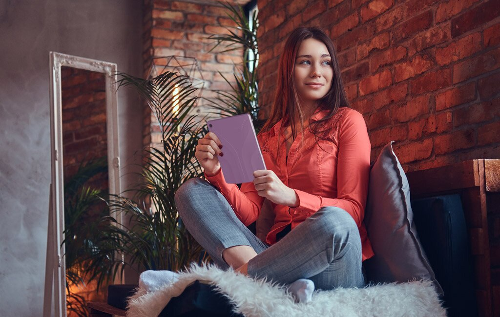 Kobieta w różowej koszuli i szarych spodniach siedzi na kanapie, trzymając tablet.