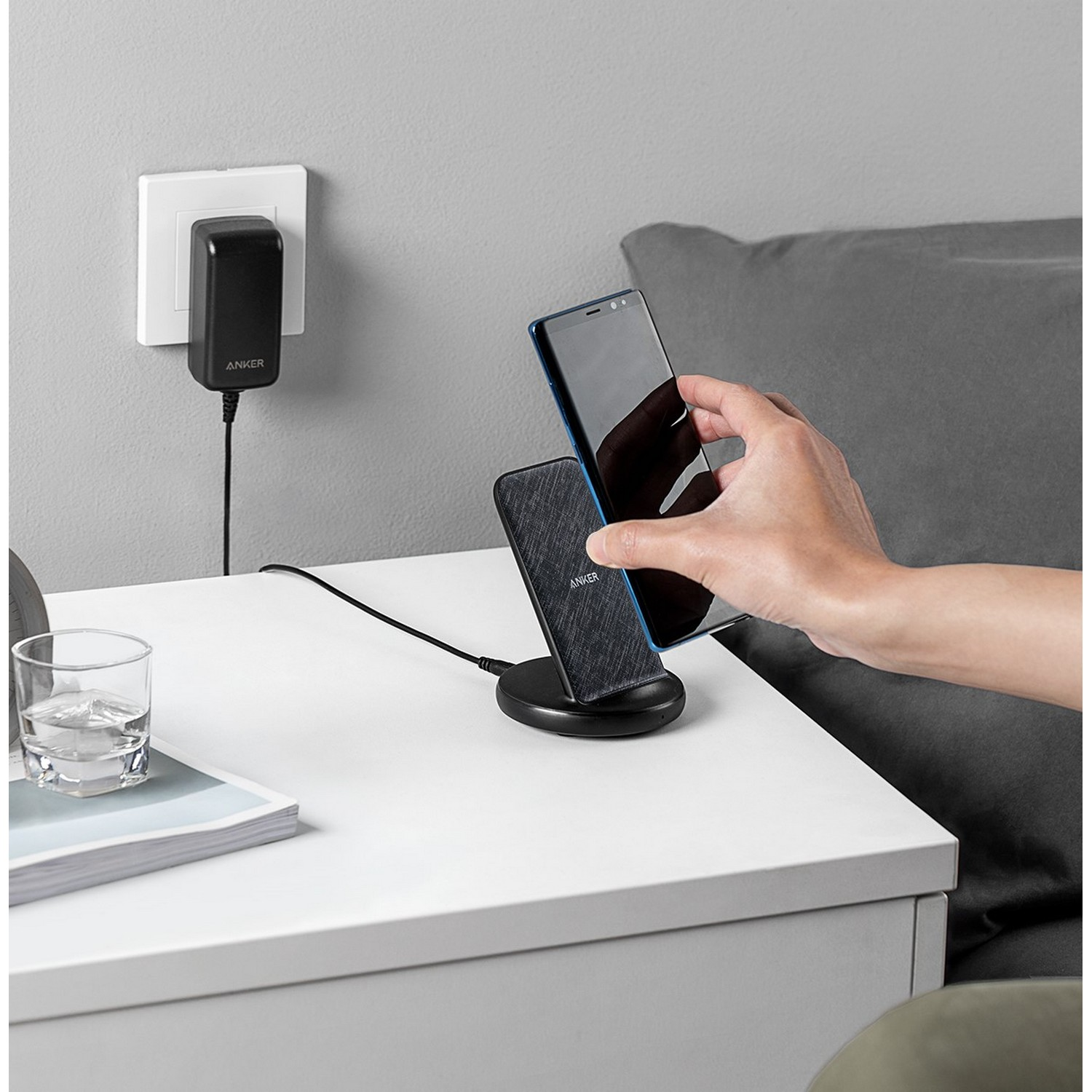 Een hand die een smartphone boven een draadloze oplader houdt, naast een wandcontactdoos en een nachtkastje.