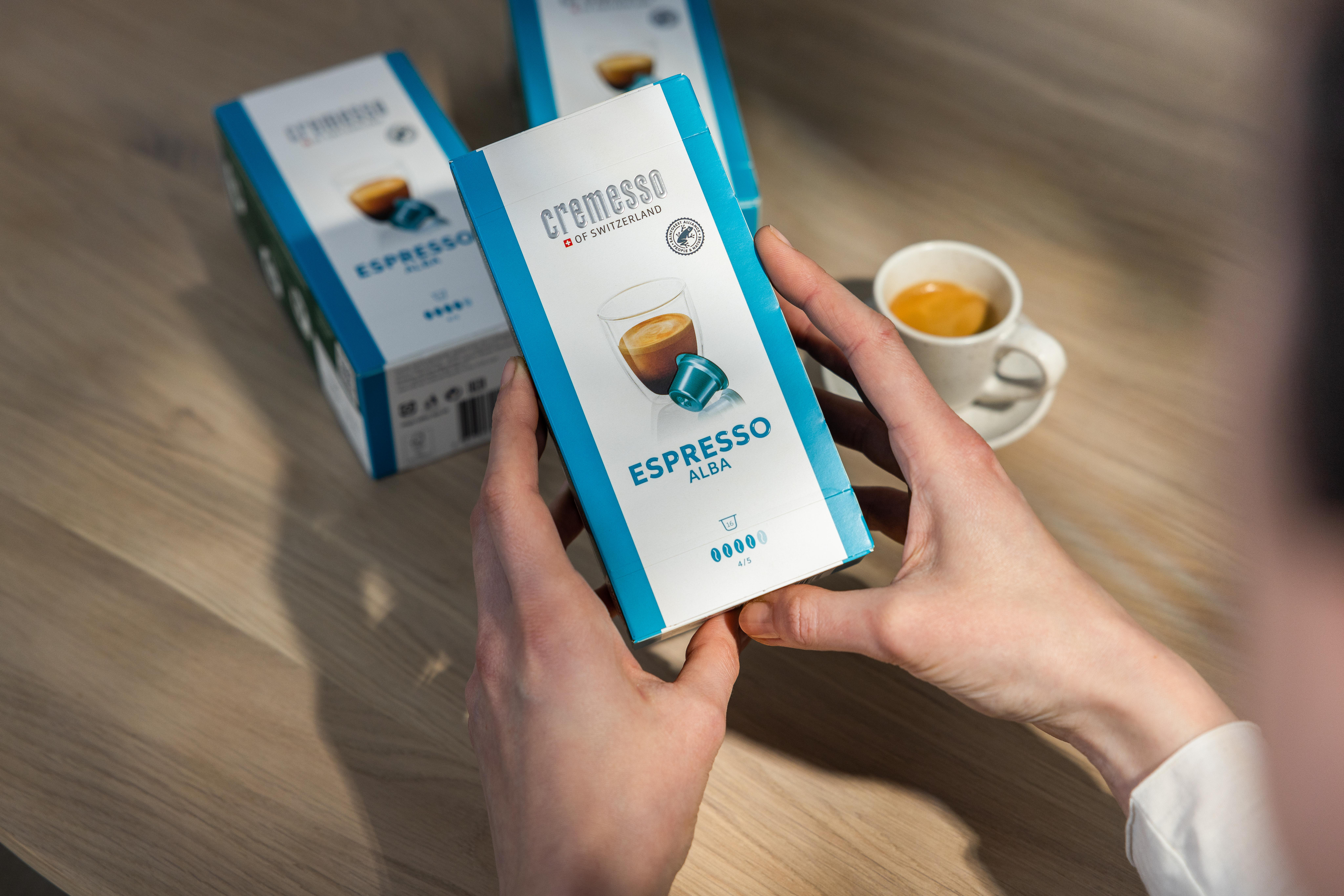 Hände halten eine Schachtel Cremesso Espresso Alba Kapseln, daneben eine Kaffeetasse.