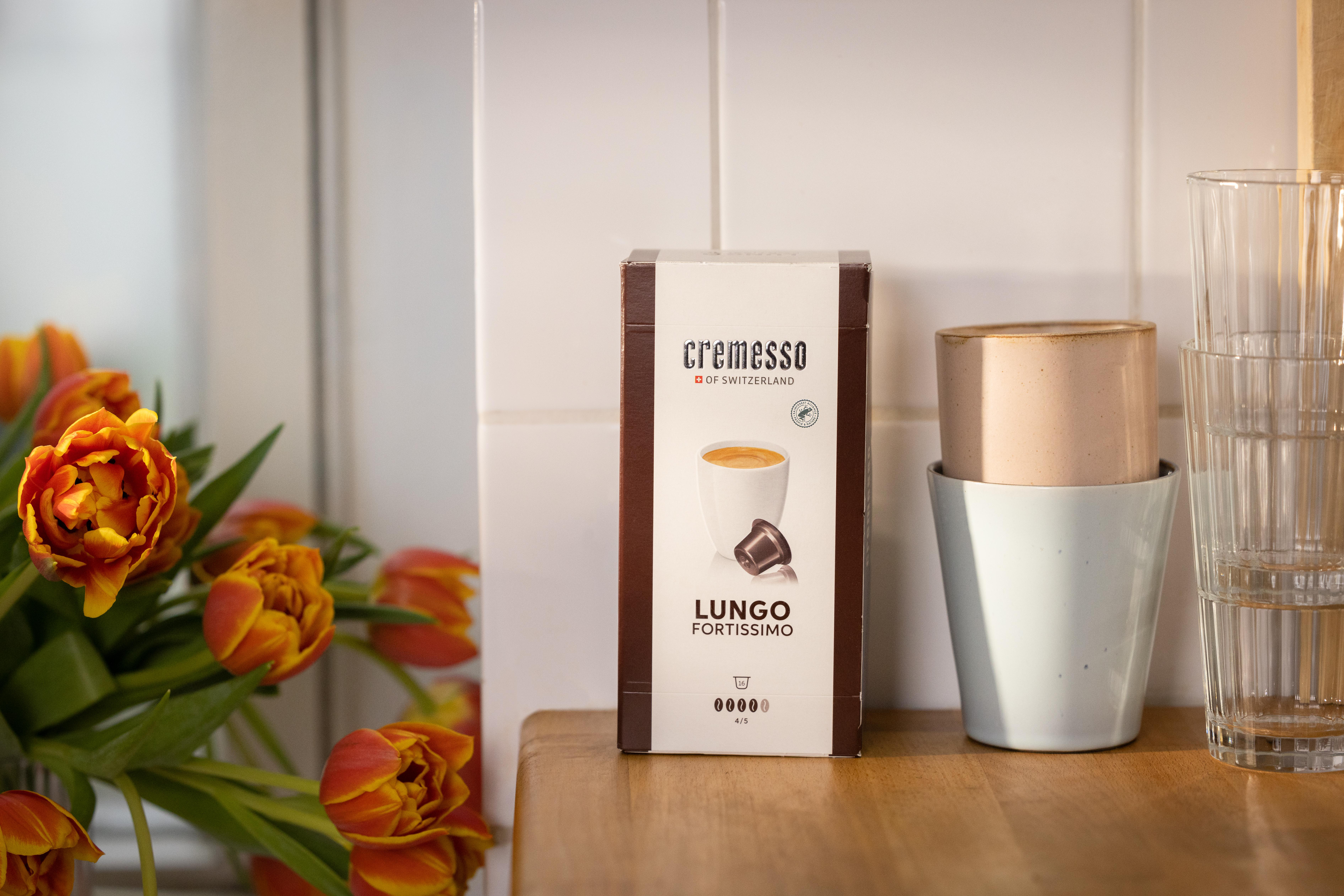 Eine Schachtel Kaffeekapseln, mit Tulpen in einer Vase und Tassen auf einem Holzregal in einer Küche.