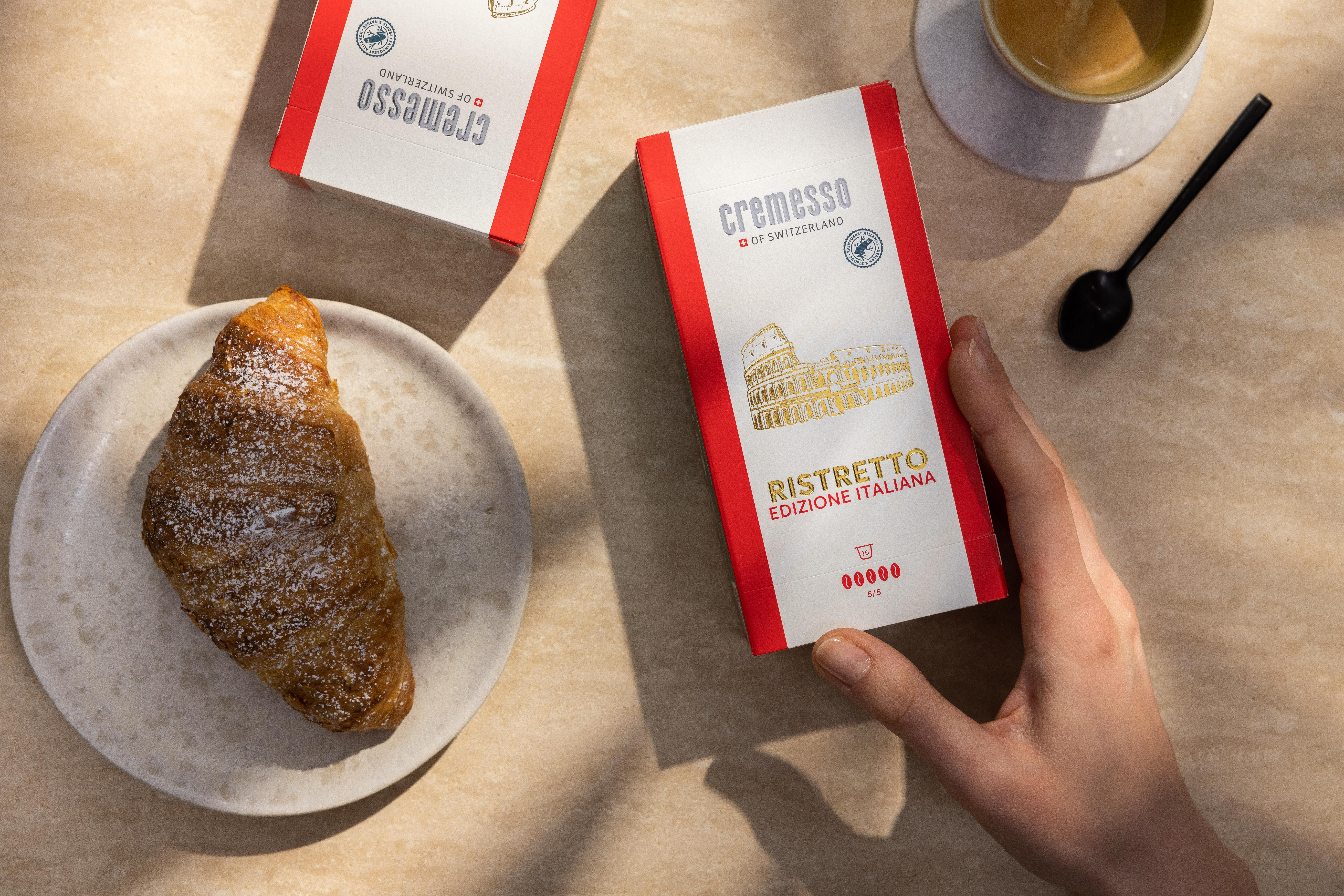 Croissant auf einem Teller, Kaffee und Kaffeepackungen. Hand greift nach einer Packung. Sonnenlicht.