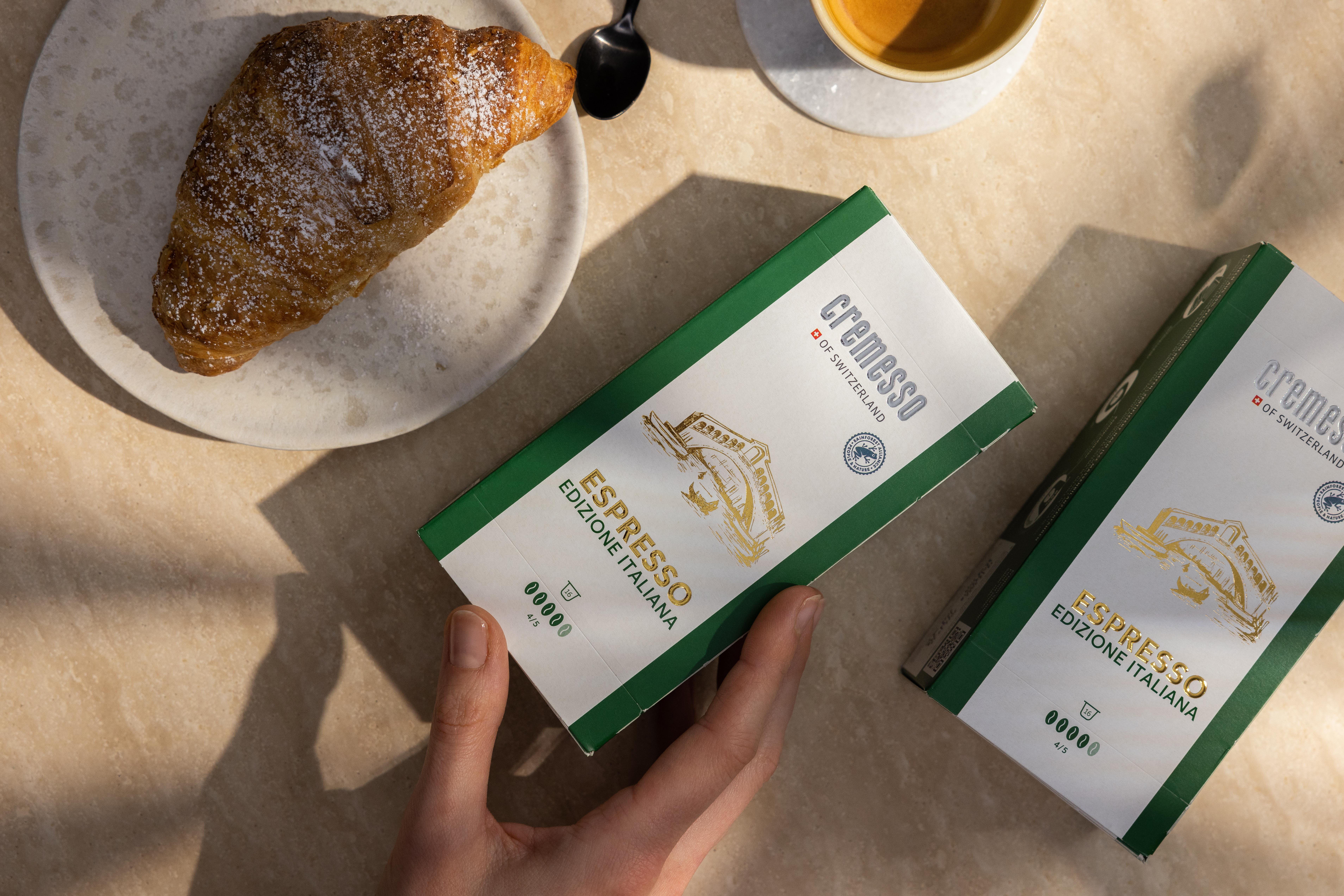Hand hält zwei Espresso-Schachteln, mit einem Croissant auf einem Teller und einer Kaffeetasse auf dem Tisch.
