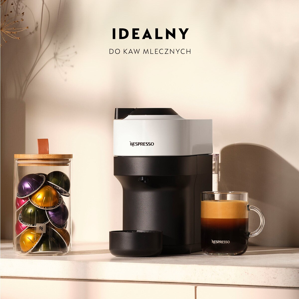 Ekspres do kawy Nespresso ze słoikiem kapsułek i filiżanką kawy na stole.