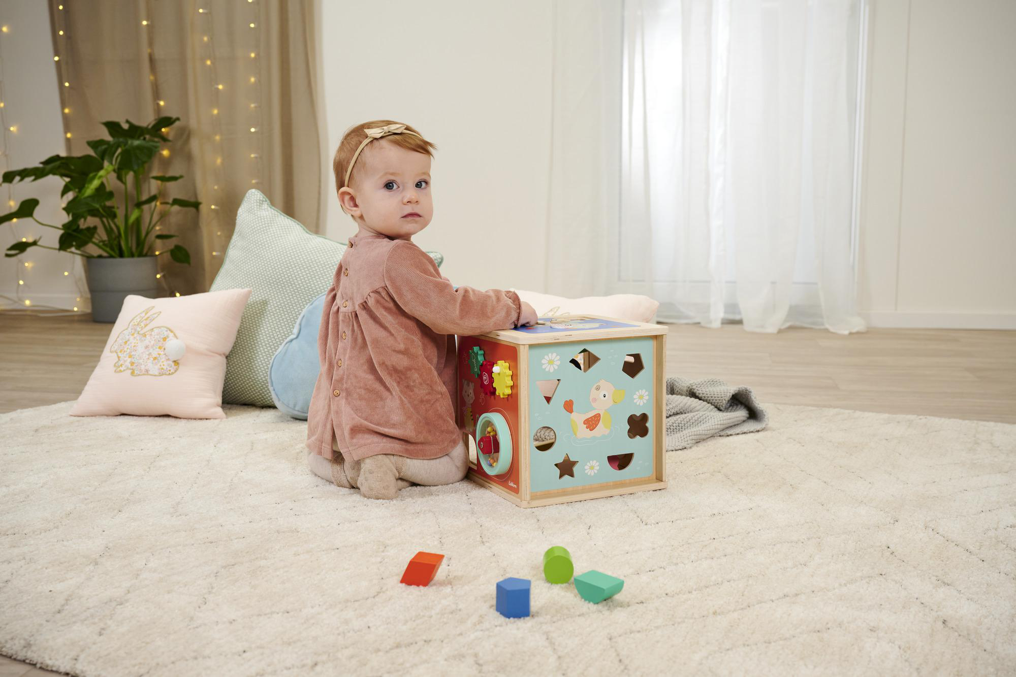 Ein Baby sitzt in der Nähe eines Holzaktivitätswürfels mit Spielzeug und einem Teppich.