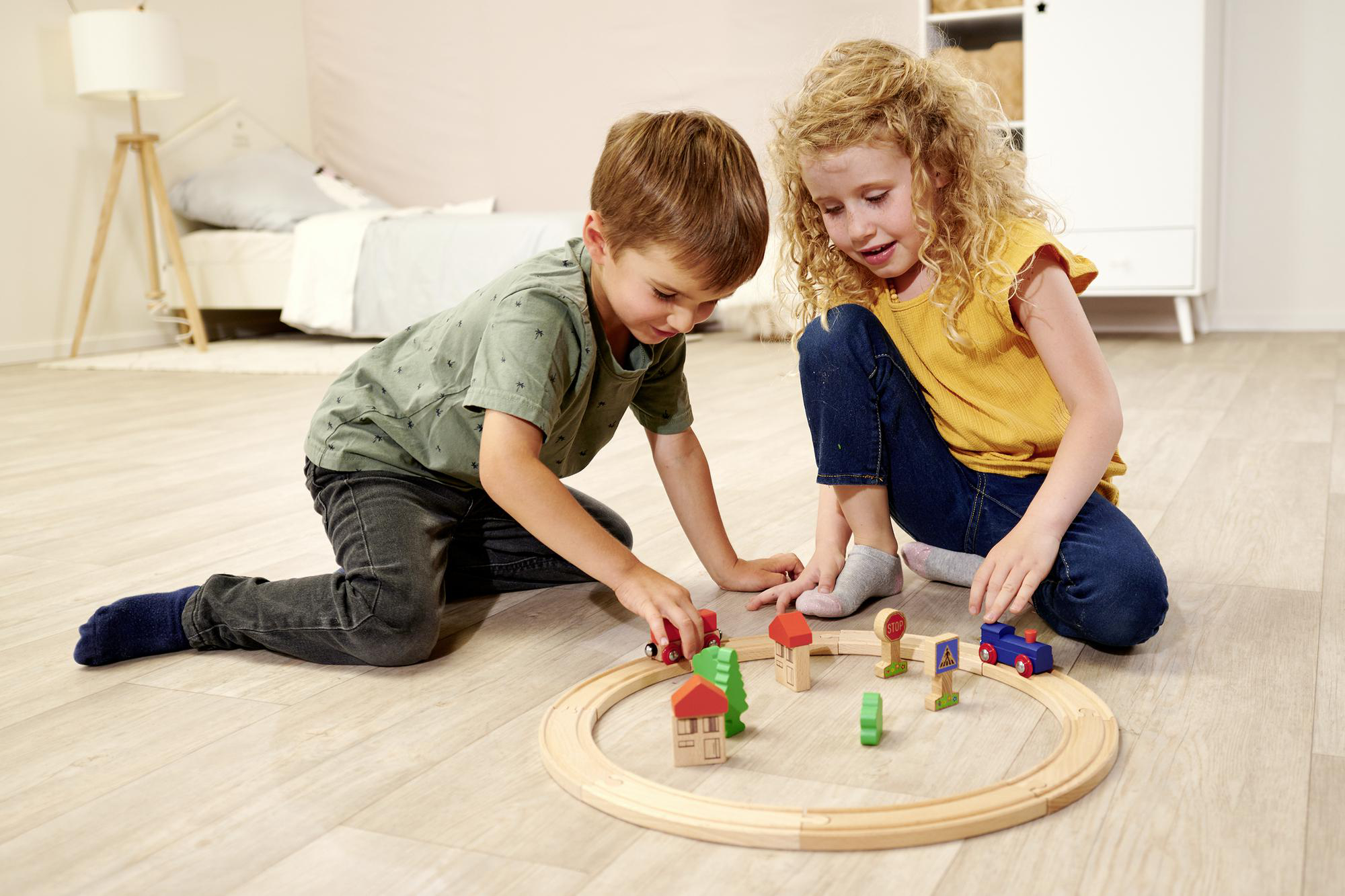 Zwei Kinder spielen mit einem hölzernen Eisenbahn-Set auf einem hellen Holzboden.