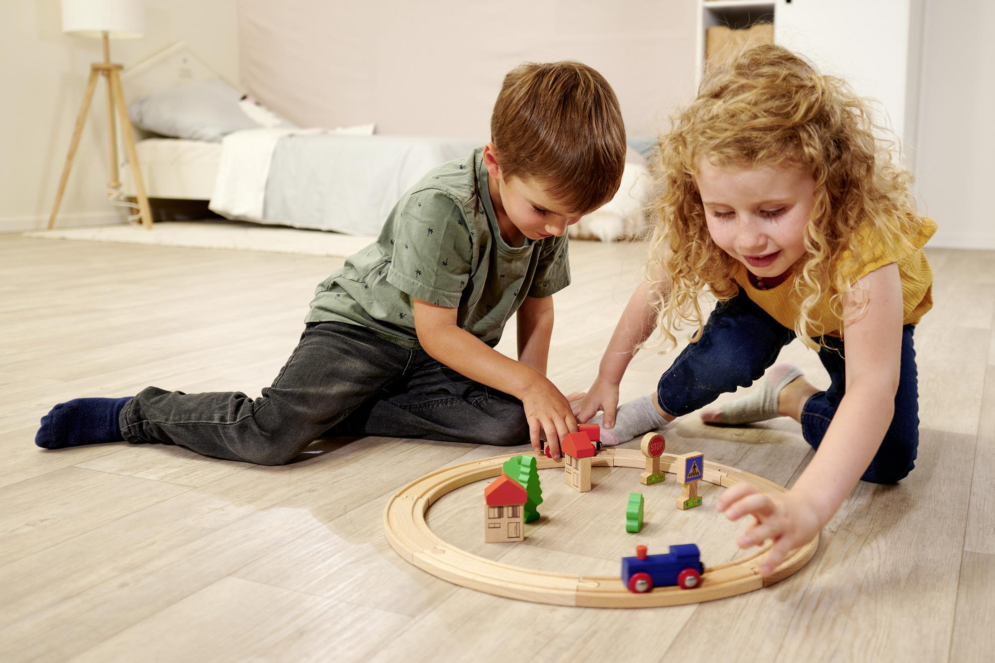 Zwei Kinder spielen mit einem Holzeisenbahn-Set auf einem Holzboden. Das Set enthält Bäume und Häuser.