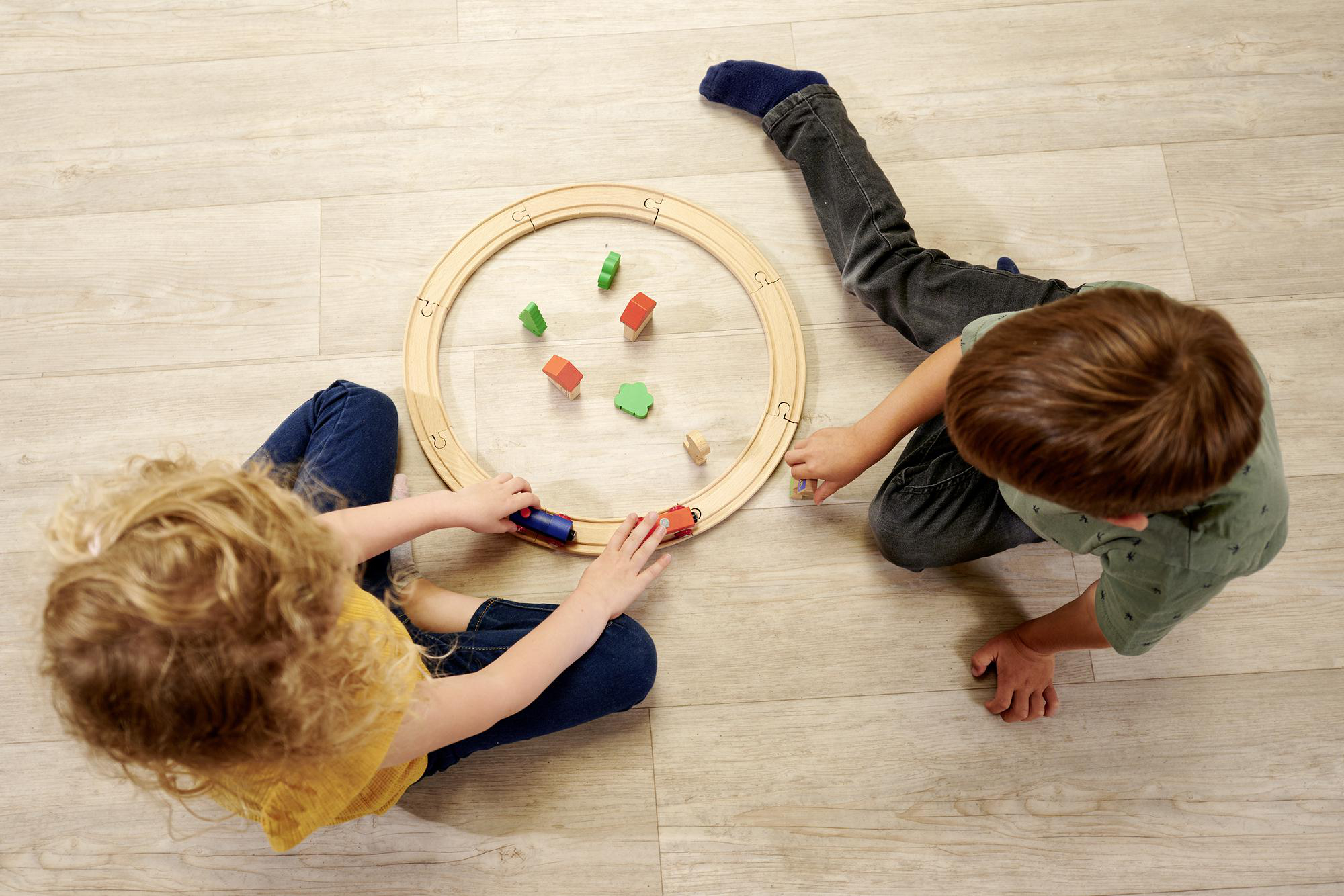 Zwei Kinder spielen mit einer Holzeisenbahn auf hellem Holzboden.