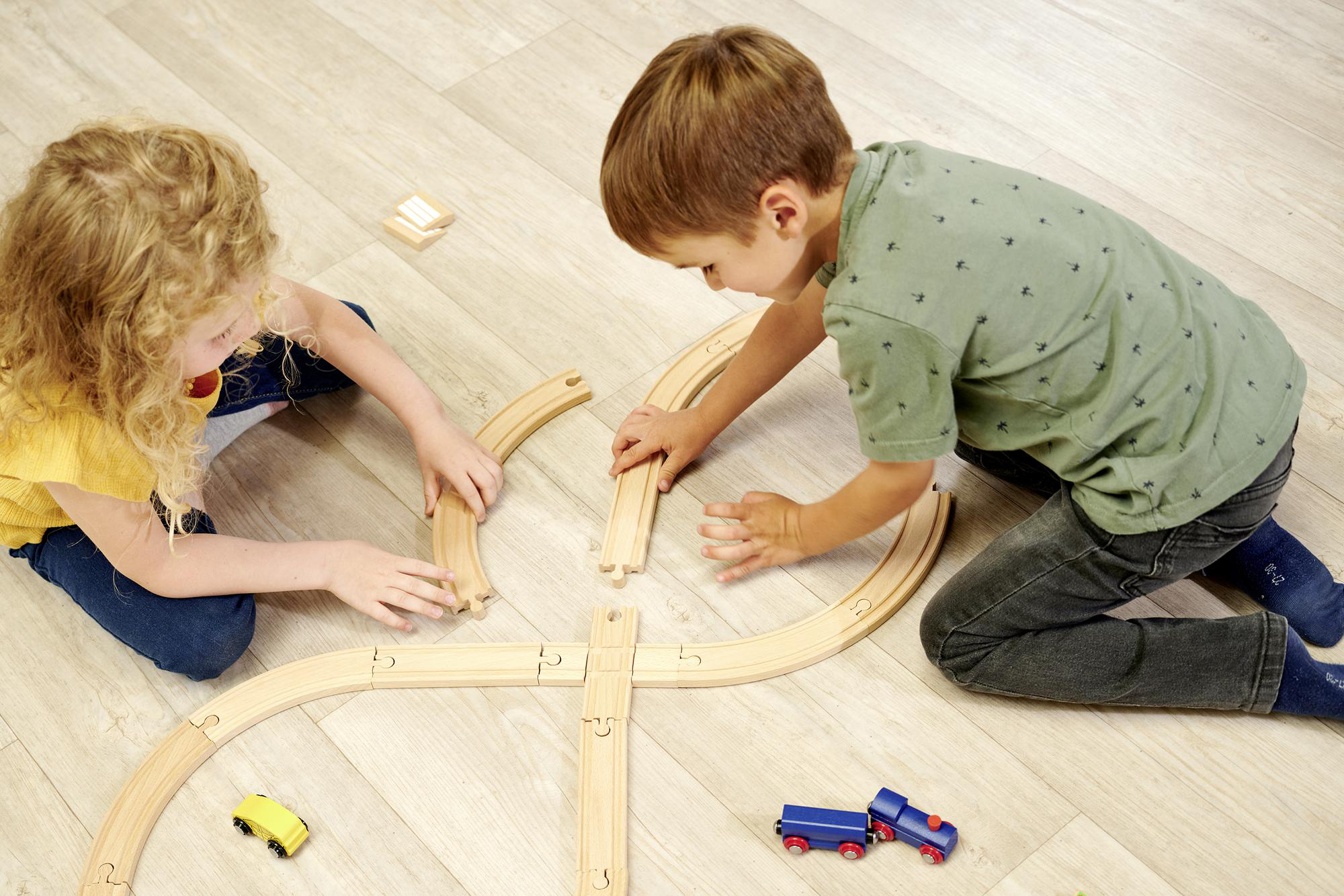 Zwei Kinder spielen mit einer hölzernen Spielzeugeisenbahn auf einem Holzboden.