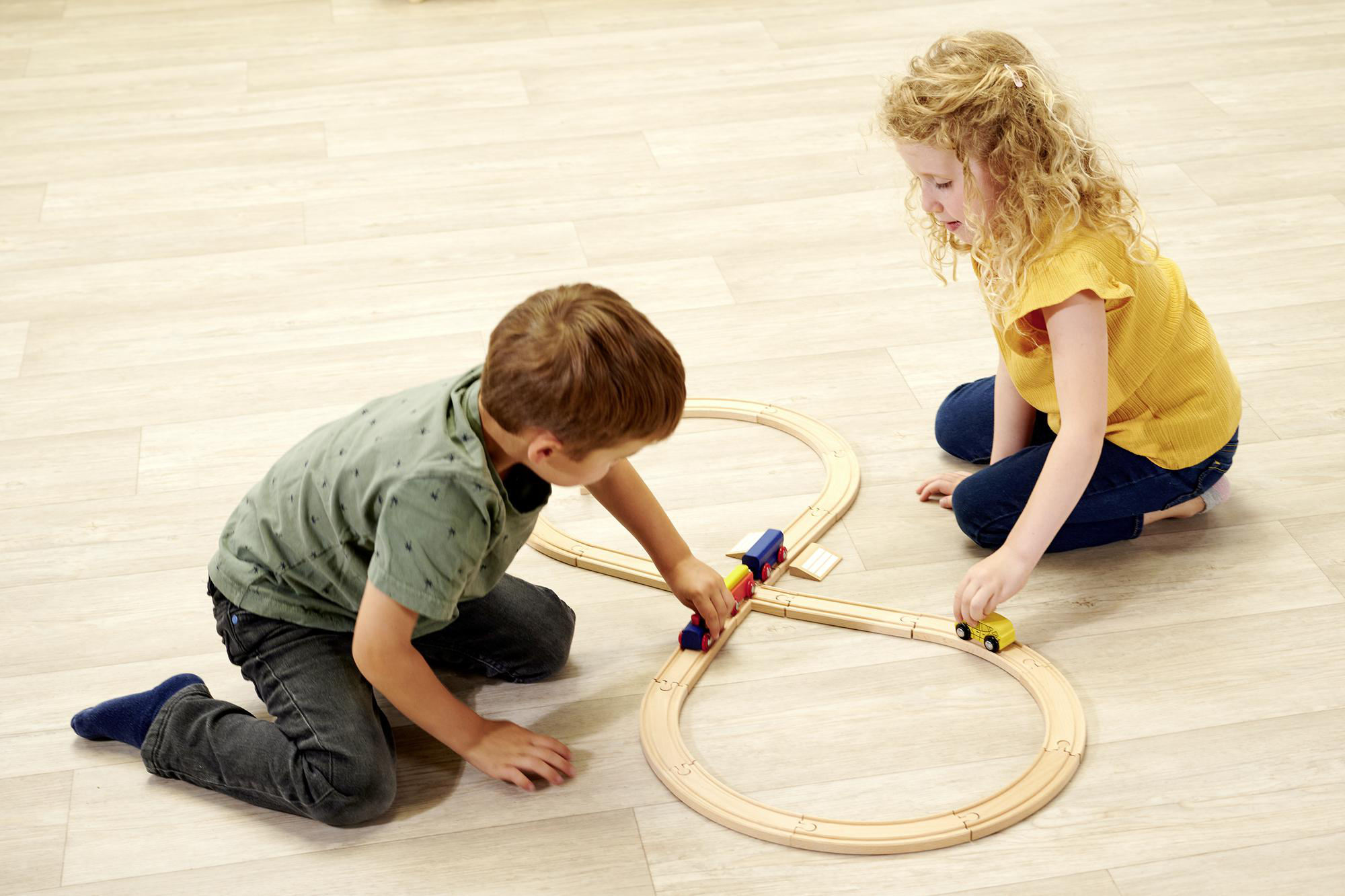 Zwei Kinder spielen mit einer Holzeisenbahn auf hellem Holzboden.
