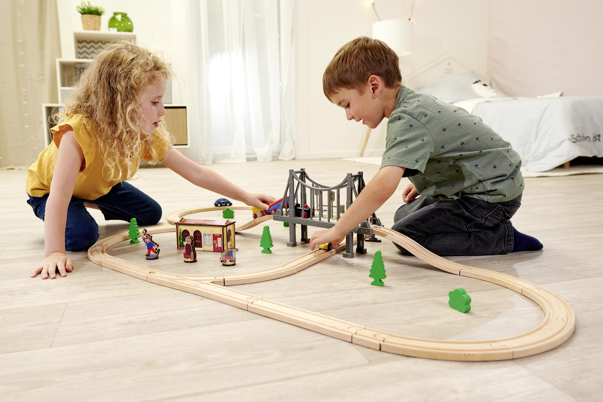 Zwei Kinder spielen mit einer Holzeisenbahn. Eine Strecke, Brücke, Züge und Bäume sind sichtbar.