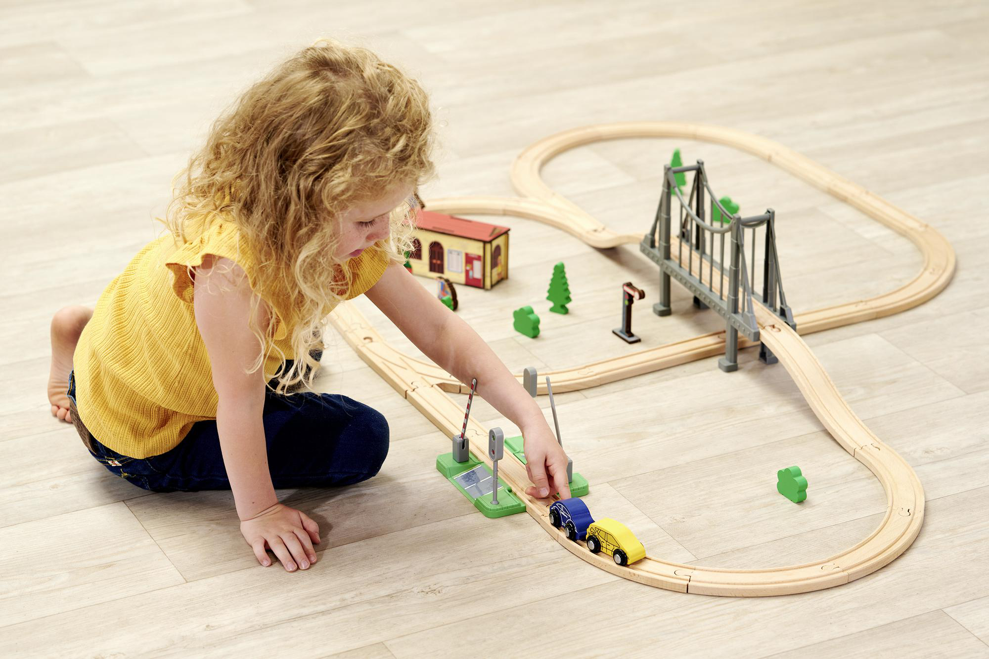 Ein junges Mädchen spielt mit einer Holzeisenbahn auf einem hellen Holzboden, mit einer Brücke und Bäumen.