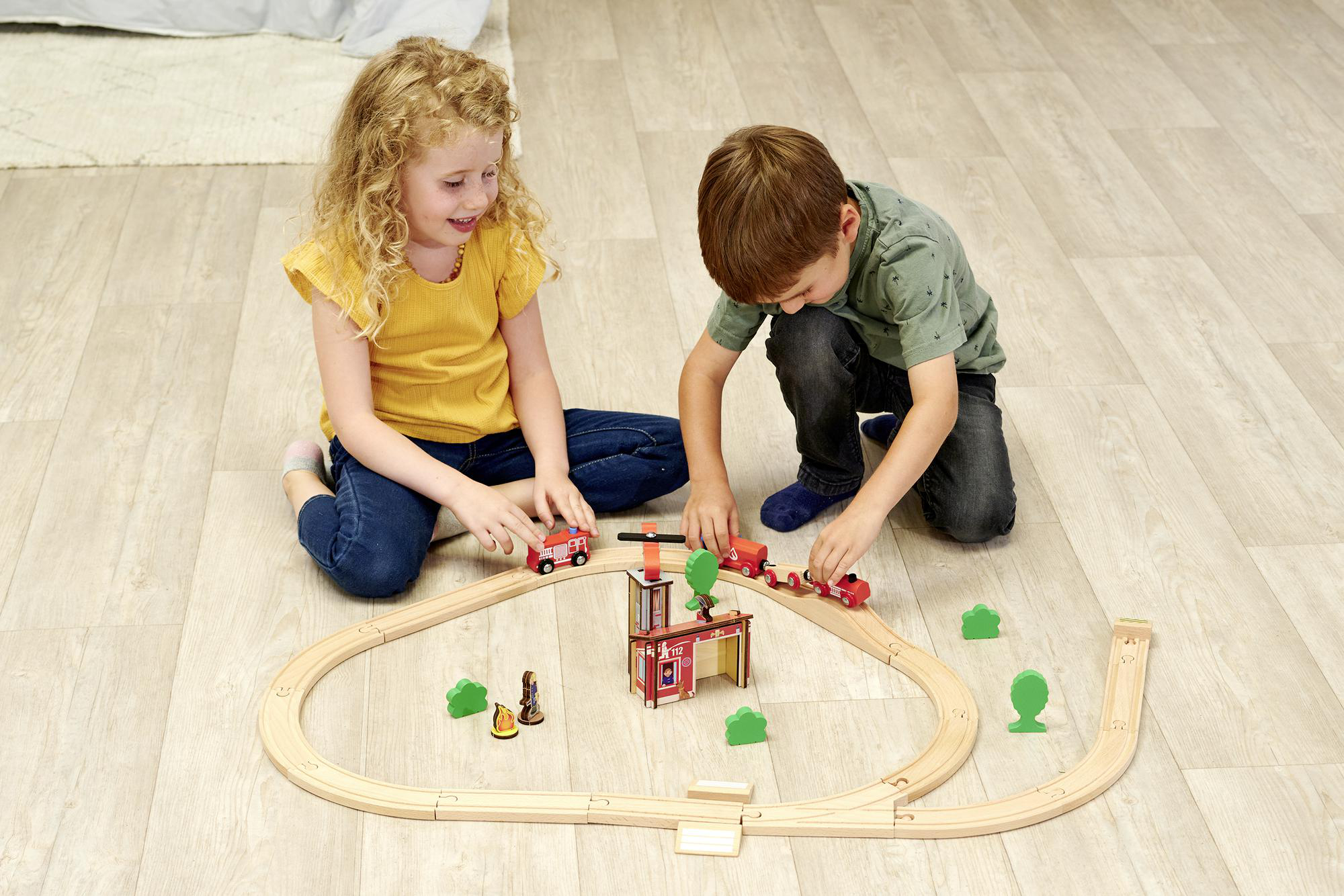 Zwei Kinder spielen mit einem Holzeisenbahn-Set mit Feuerwache und Feuerwehrautos.