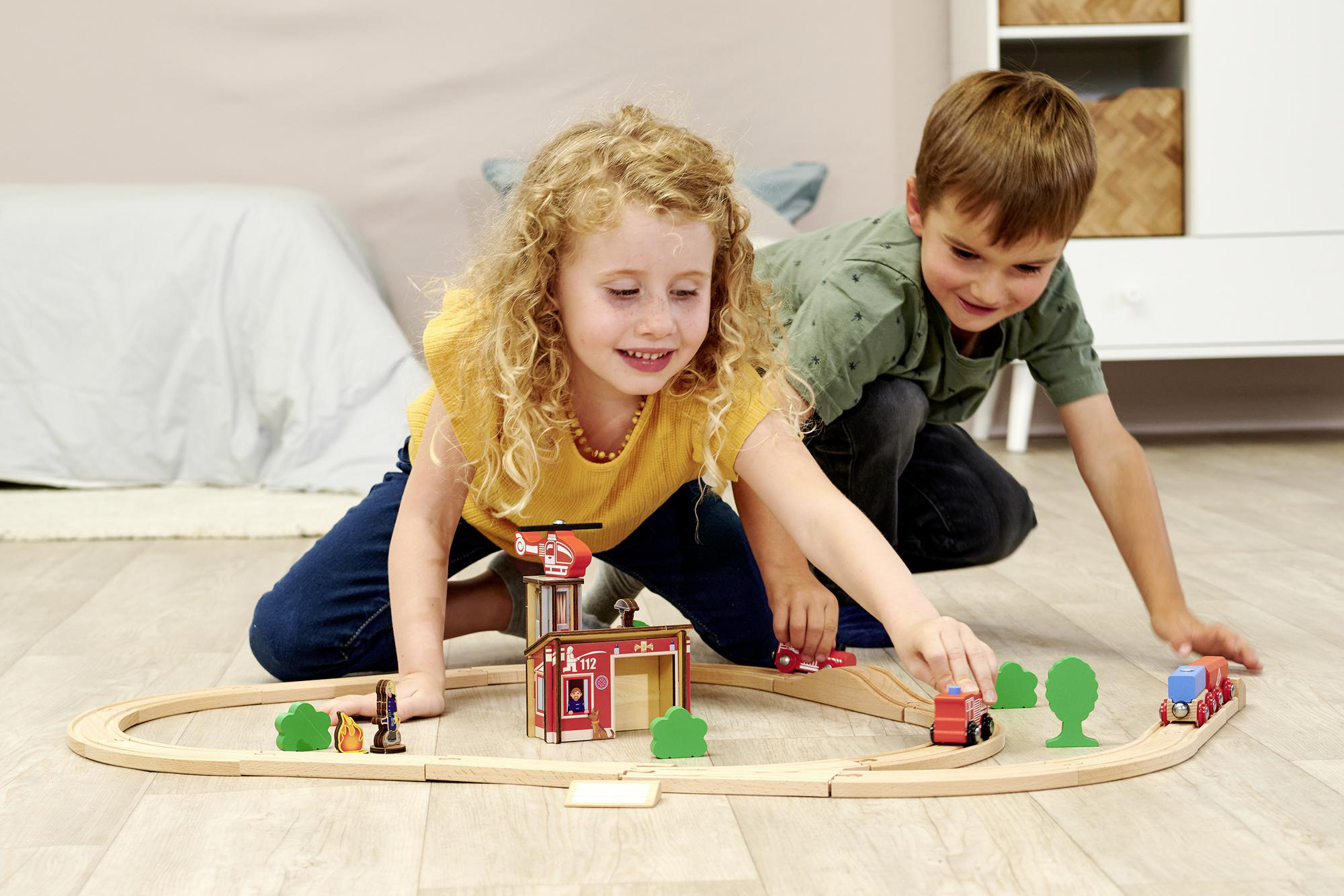 Zwei Kinder spielen mit einem hölzernen Eisenbahnset mit Feuerwache und Zug.