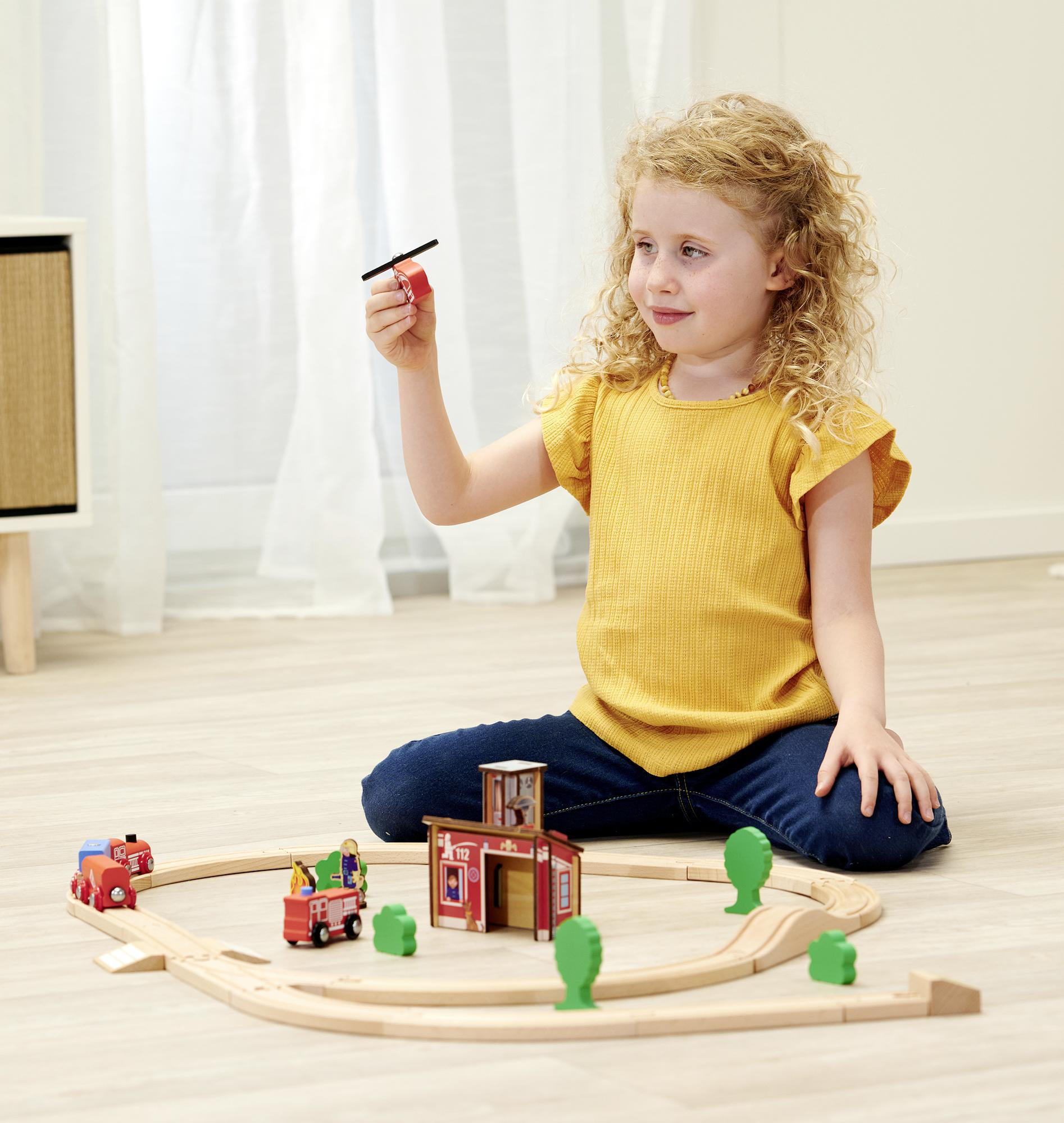 Ein junges Mädchen spielt mit einem Holzspielzeug-Eisenbahn-Set und hält ein Hubschrauberspielzeug. Das Set enthält einen Zug und Gebäude.