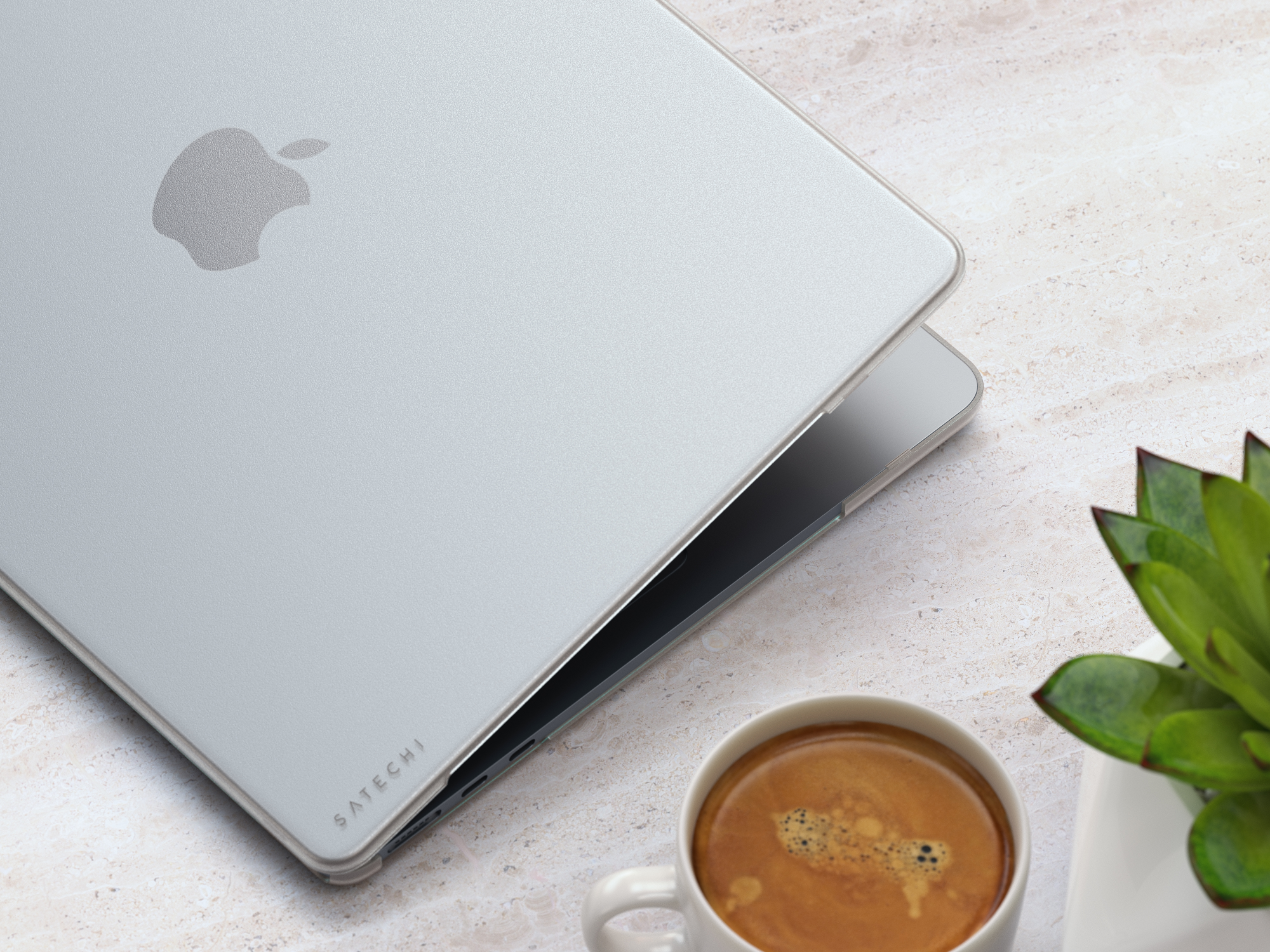Custodia per laptop Apple argentata su un tavolo con una tazza di caffè e una pianta in un vaso bianco.