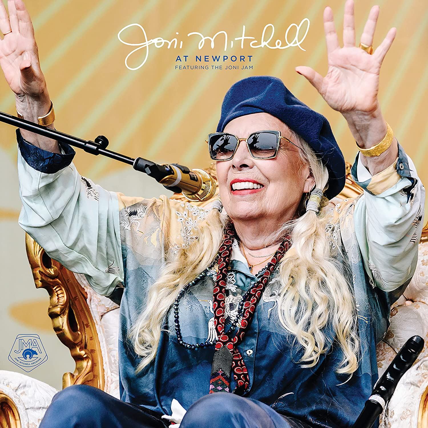 Joni Mitchell egy koncerten énekel, karjai felfelé. Napszemüveget, kalapot és mintás inget visel.