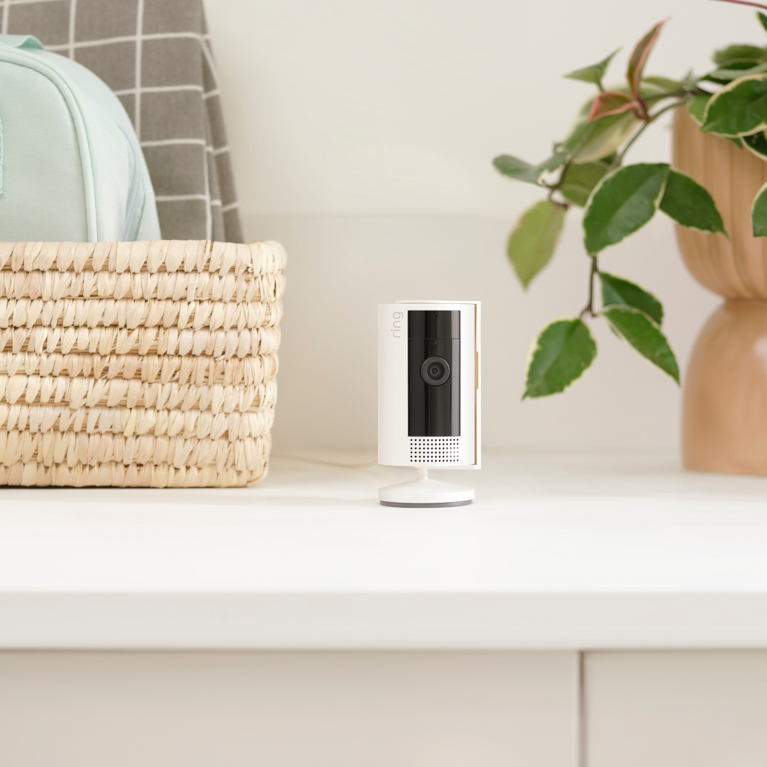 A white Ring security camera on a white surface, with a basket and plant in the background.