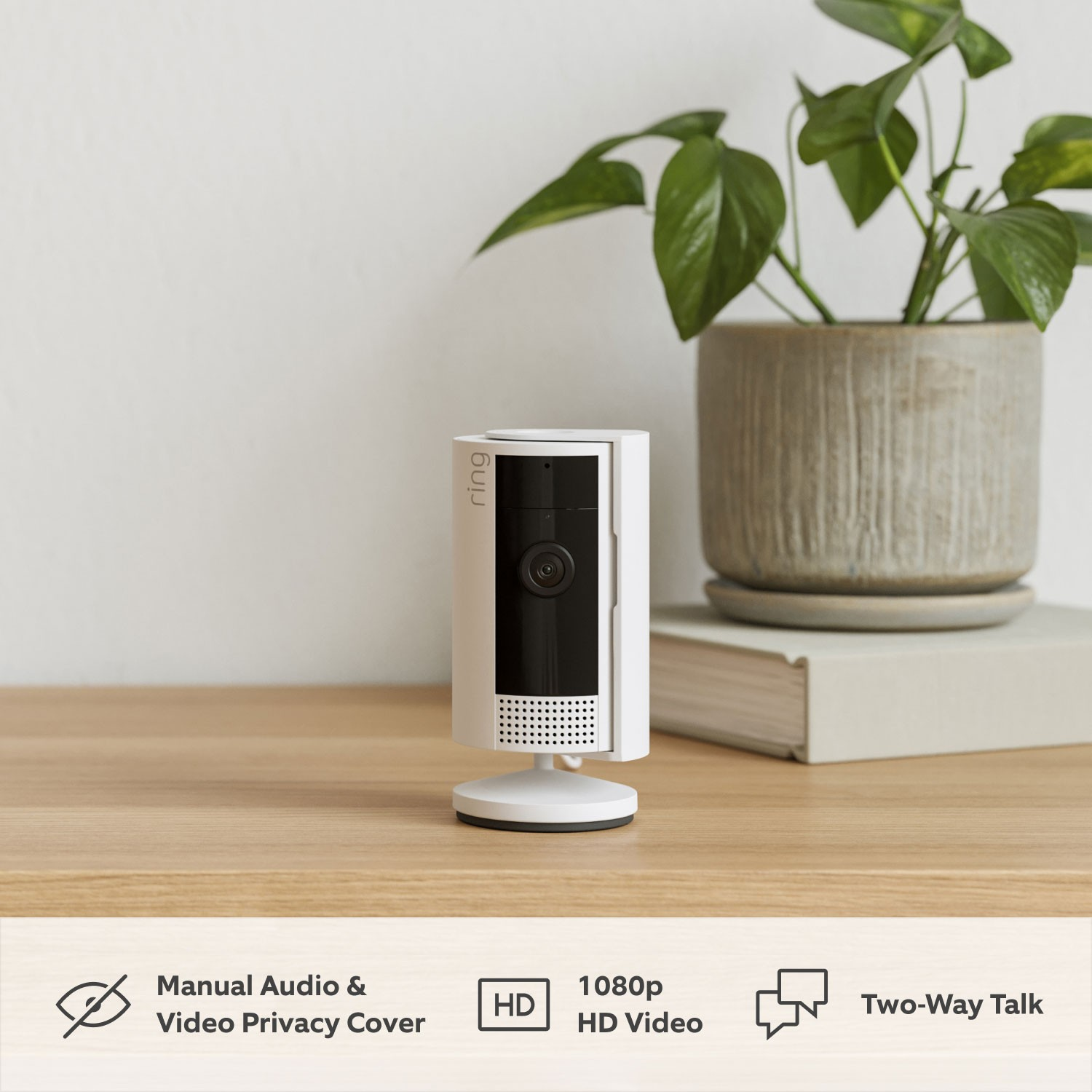 White Ring security camera. It has a black lens, is on a wooden table, and there is a plant in the background.