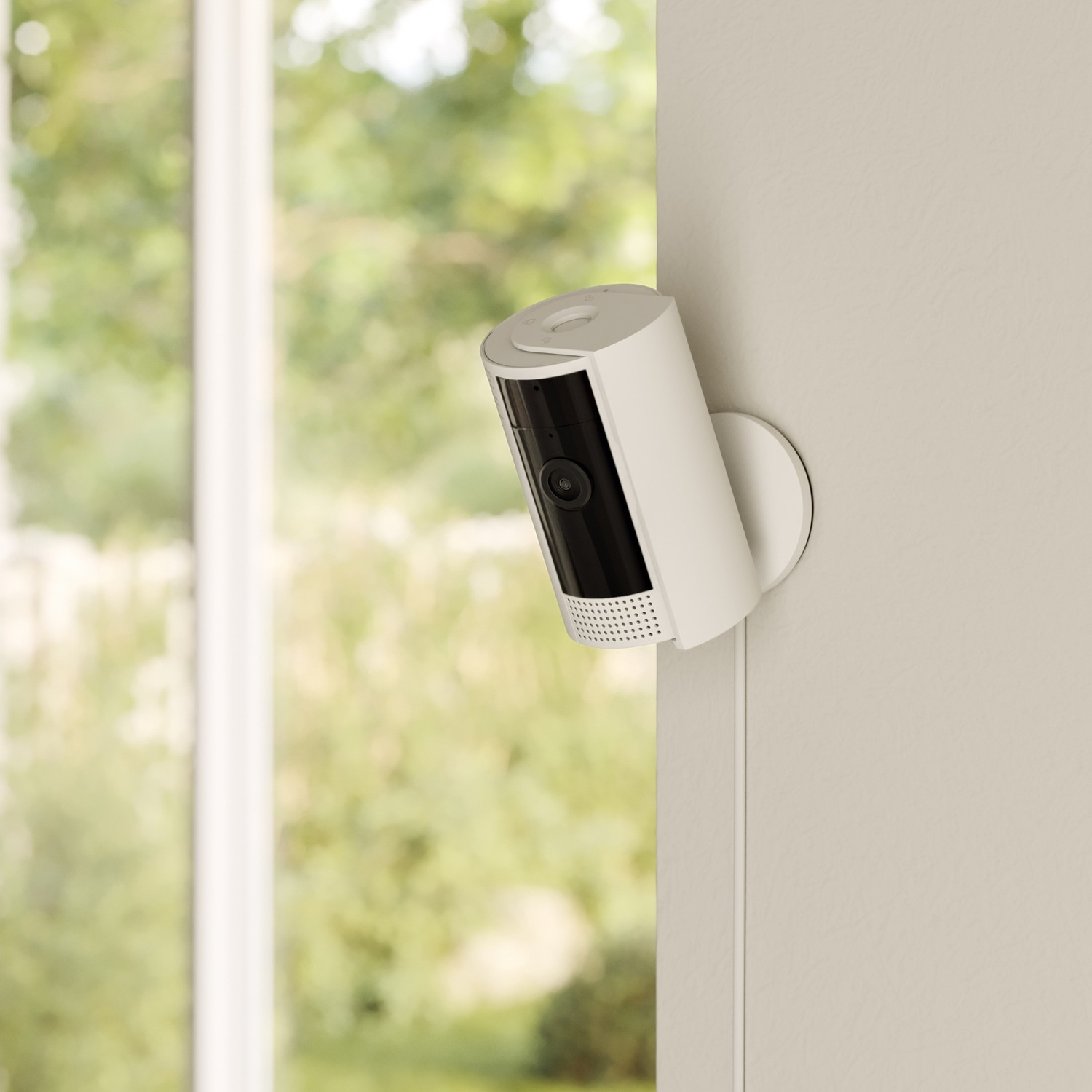 A white security camera is mounted on a wall near a window with trees visible outside.