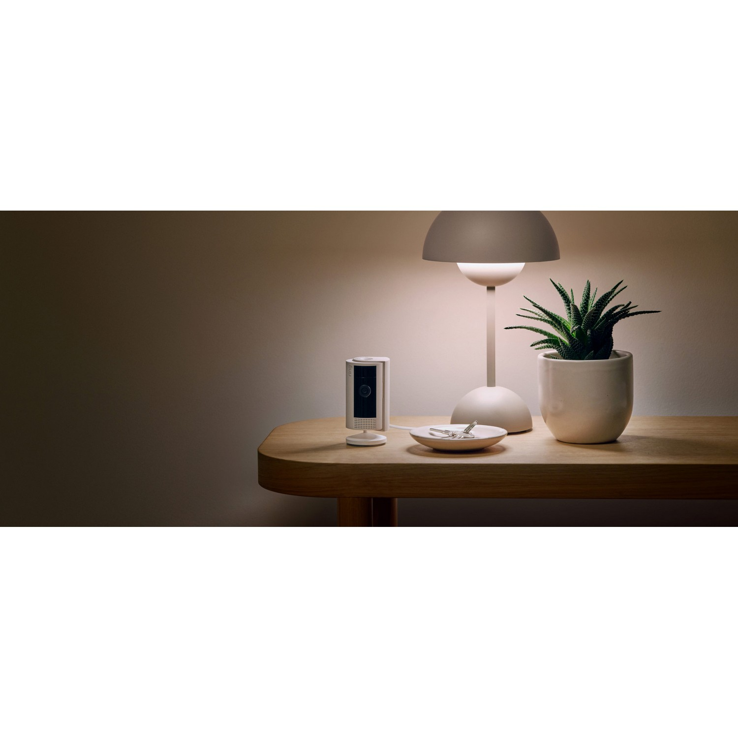 Une caméra de sécurité Ring sur une table avec une lampe et une plante. Des clés dans une petite coupelle.