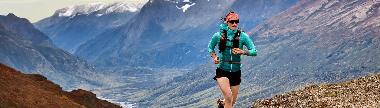 Coureur en veste turquoise et short noir courant sur un sentier de montagne.