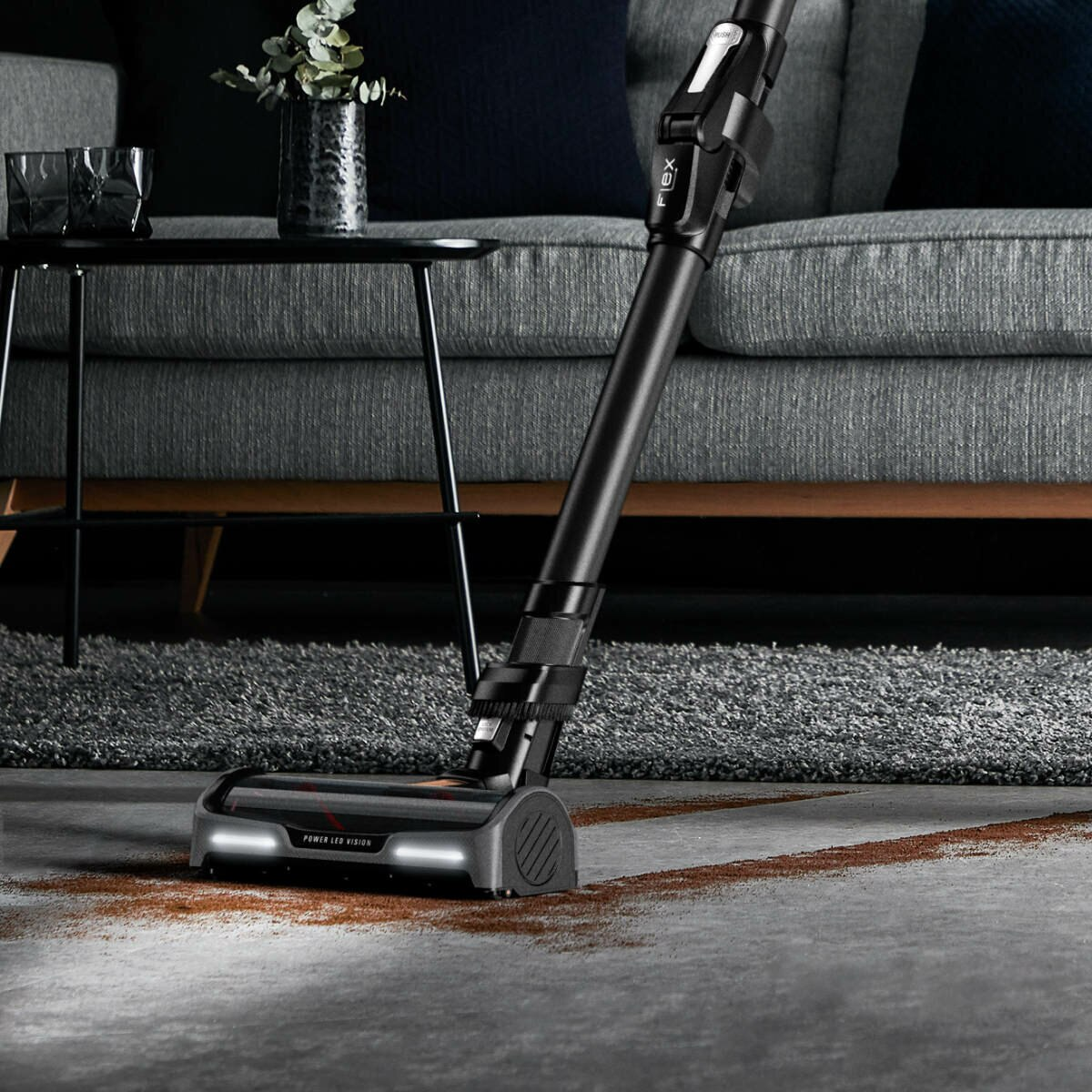 Odkurzacz na dywanie z podświetlonymi światłami, szara sofa w tle.