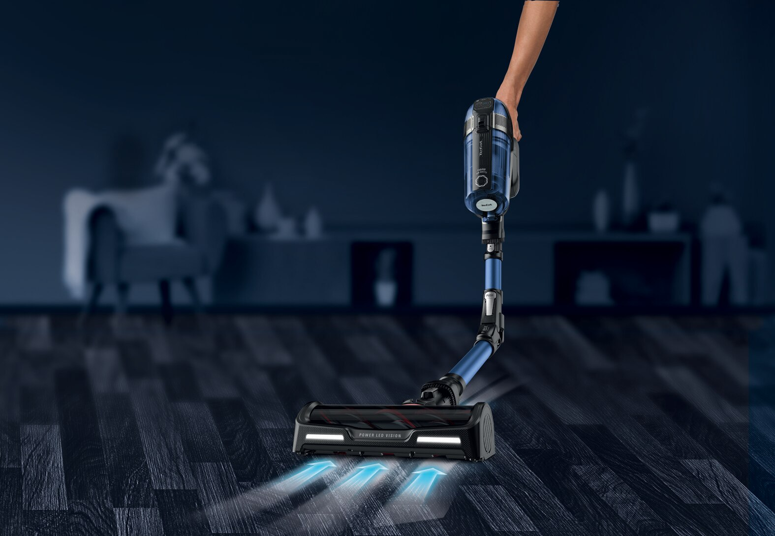 Osoba trzymająca niebiesko-czarny odkurzacz, pokazująca podłogę i dom.