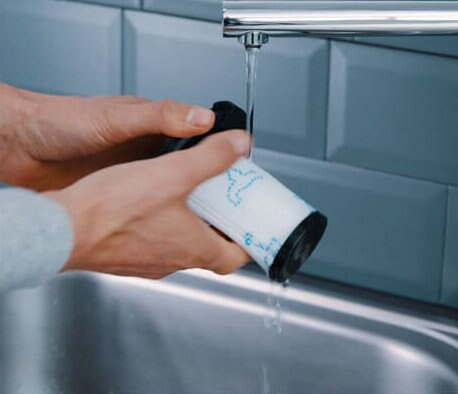 Dłonie myjące filtr od odkurzacza pod bieżącą wodą. Zlew w tle.