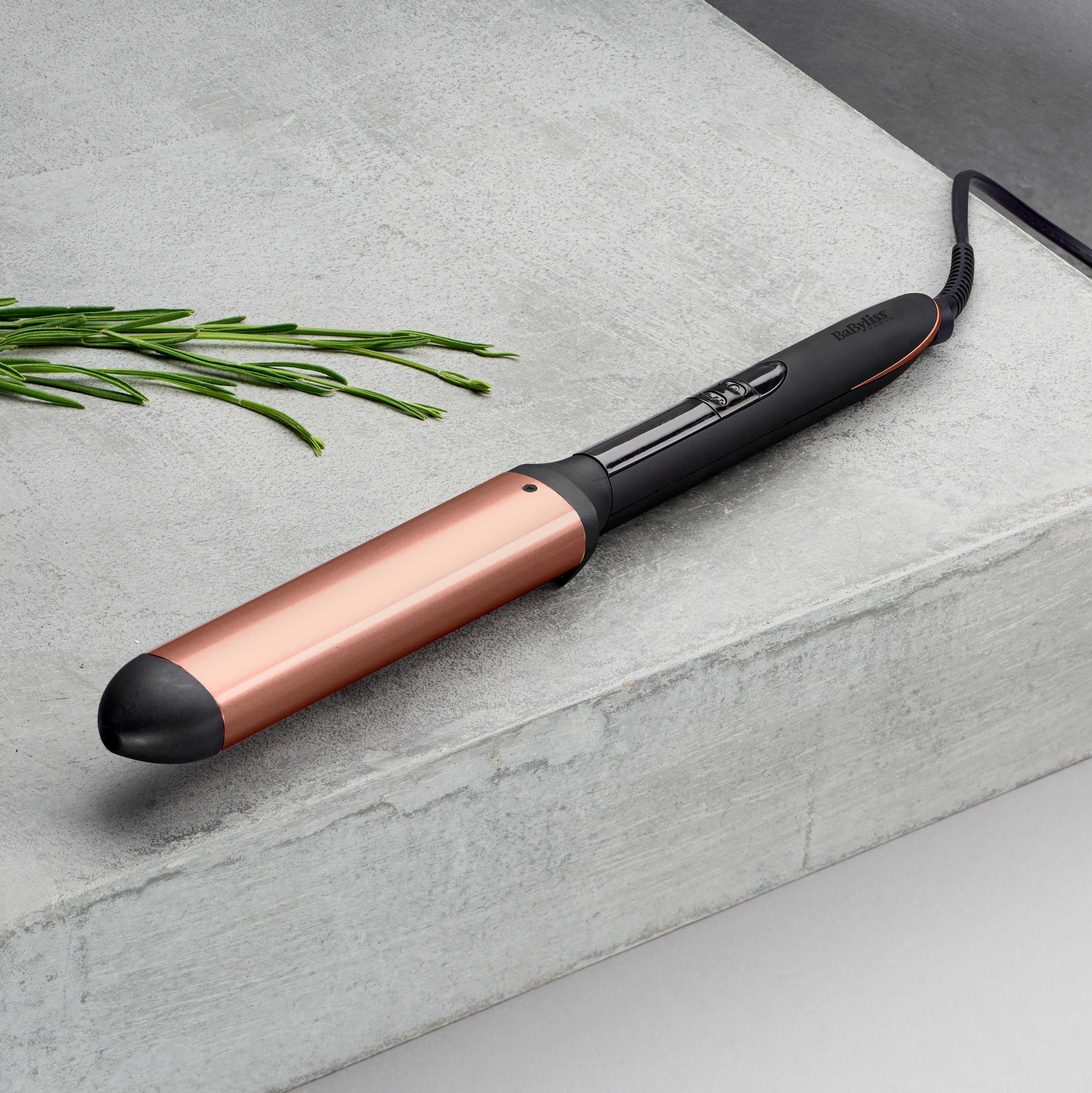 Ein Lockenstab in Schwarz und Roségold auf grauer Oberfläche mit Rosmarin.