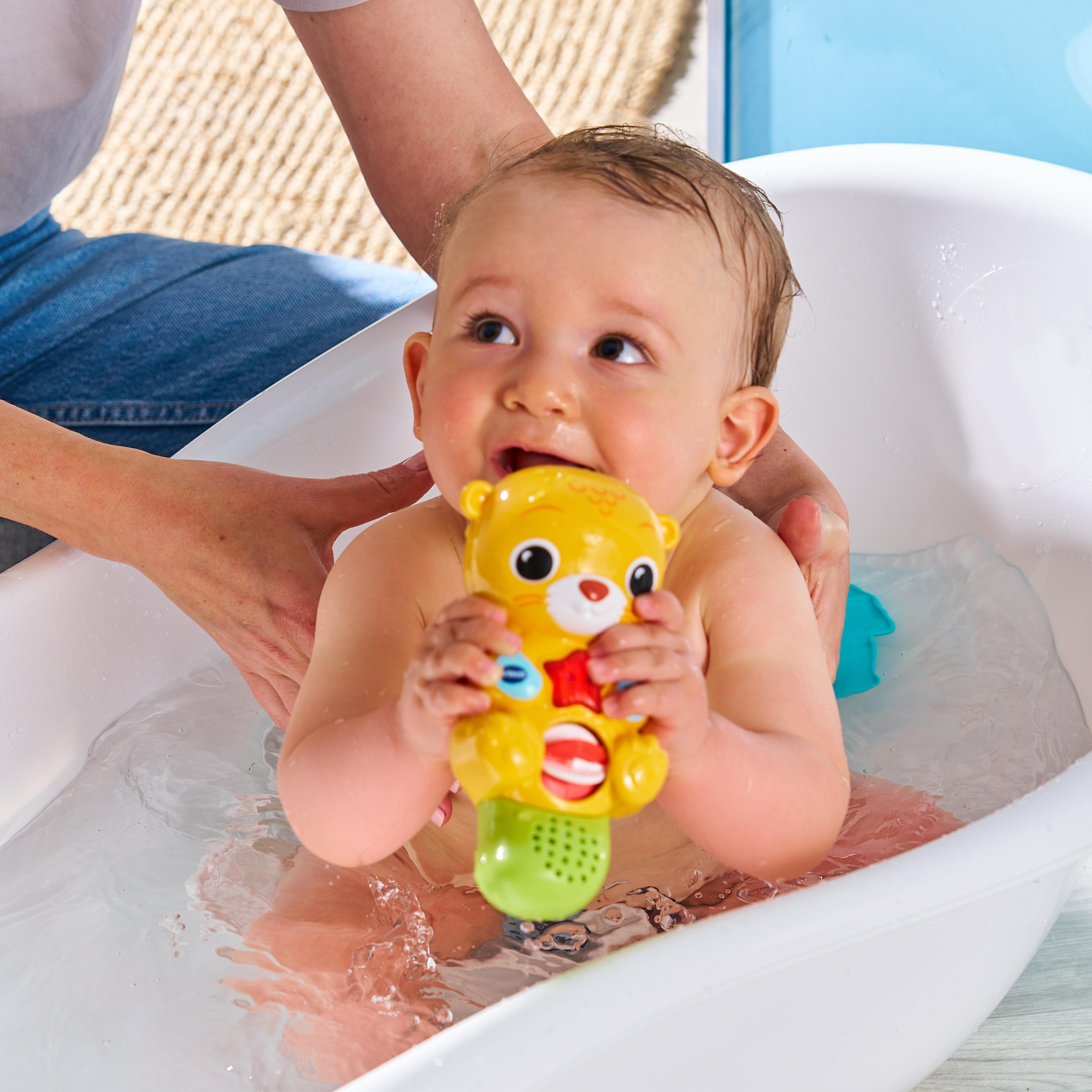 Baby in einer weißen Wanne, das ein gelbes Spielzeug hält, Hände einer Person im Blick.